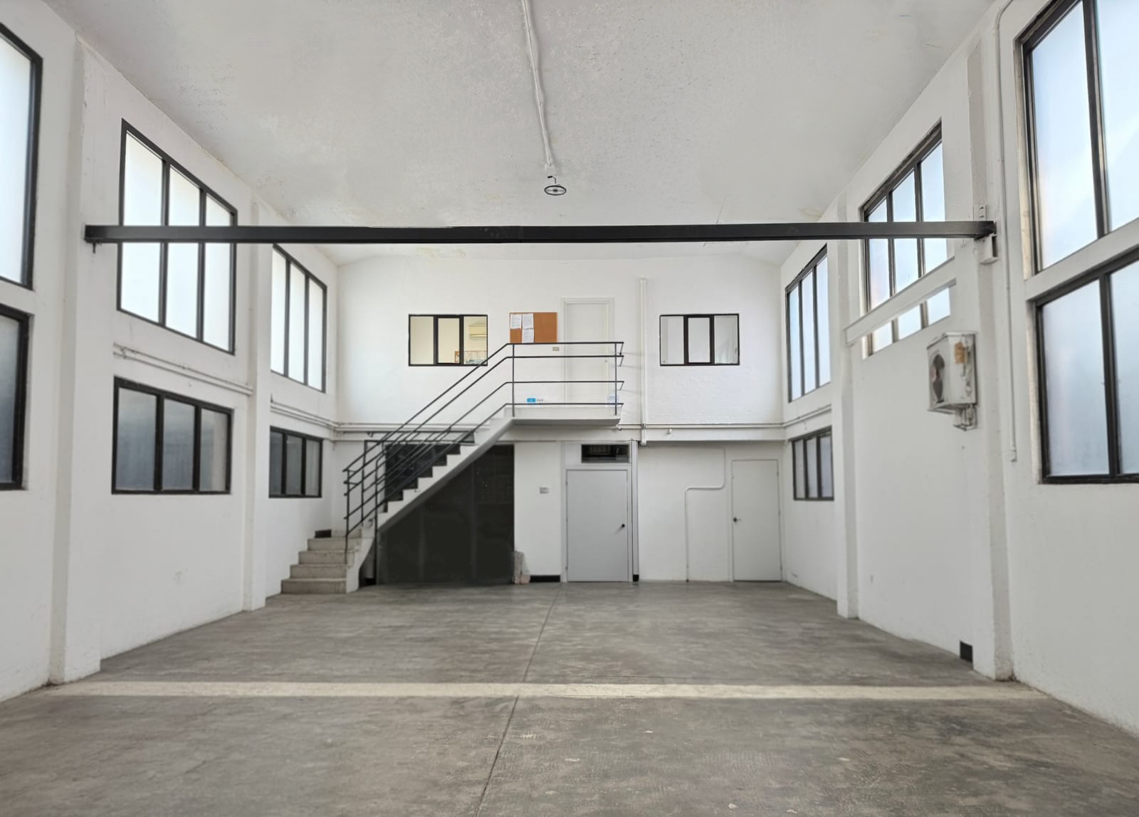 Interno di un edificio industriale o commerciale con pareti bianche, grandi finestre e pavimento in cemento, una scala metallica conduce al piano superiore.