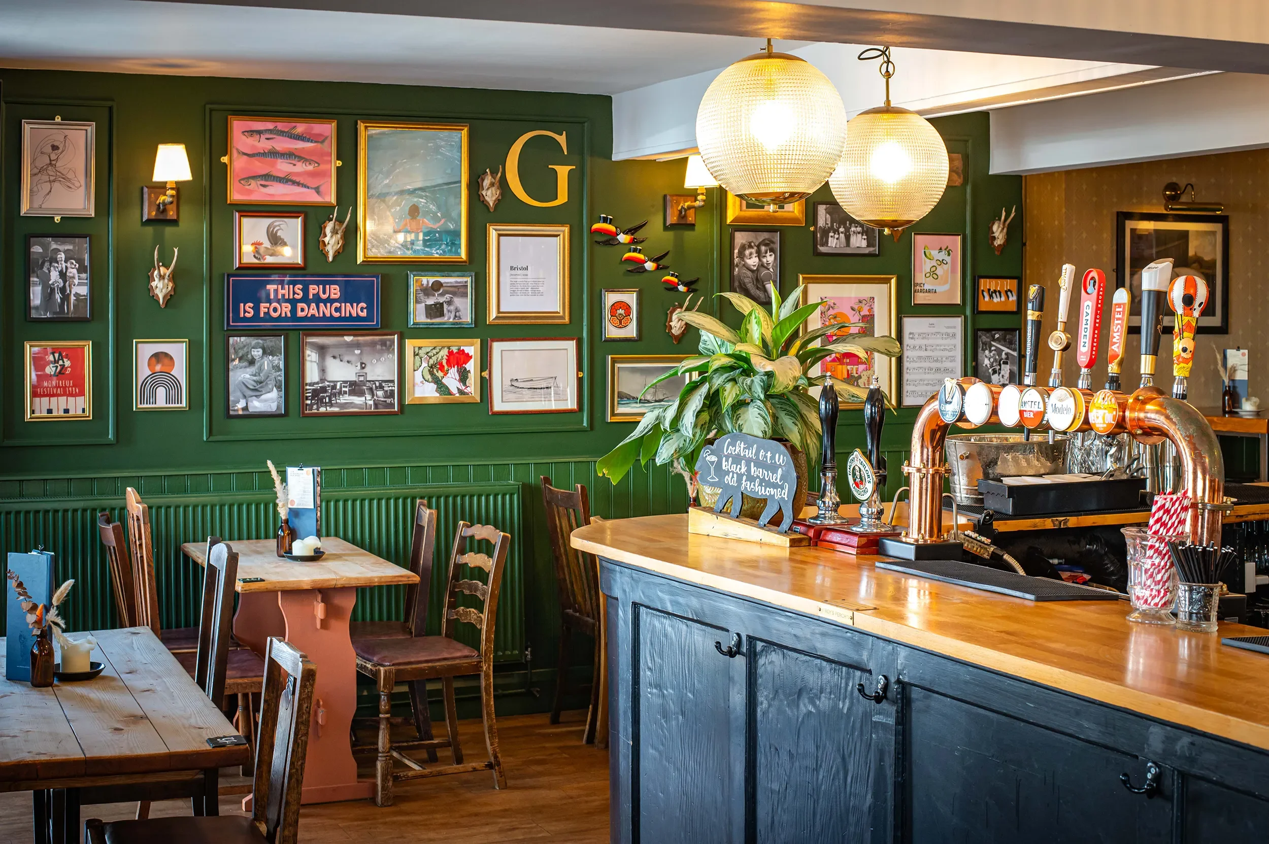Cosy pub dining area with wooden tables and chairs next to an eclectic green gallery wall with framed pictures and vintage decor.
