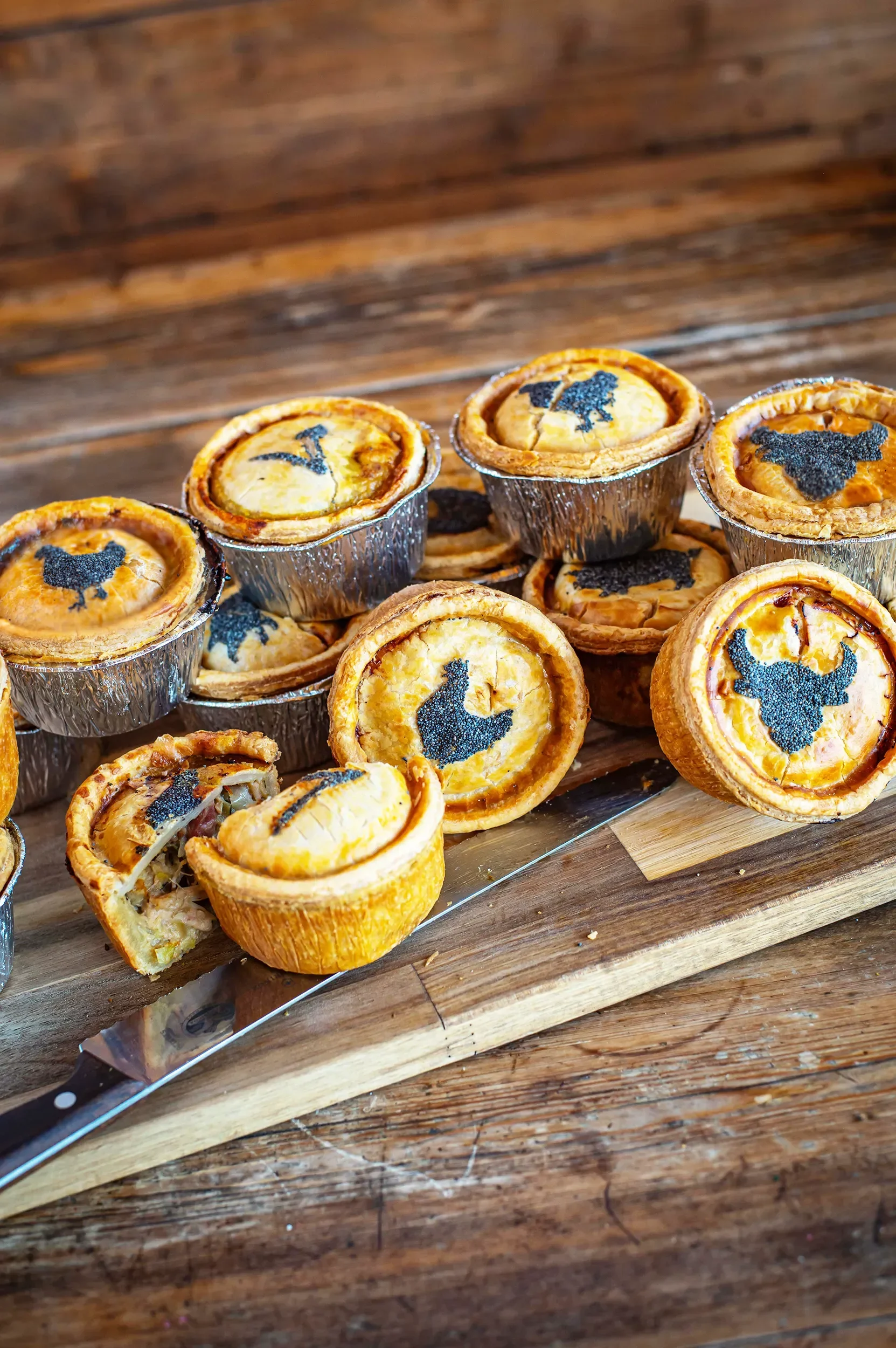 An assortment of individual artisan pies in foil tins showing chicken, beef, and vegan stencils, with one pie cut open to showcase the savory filling.