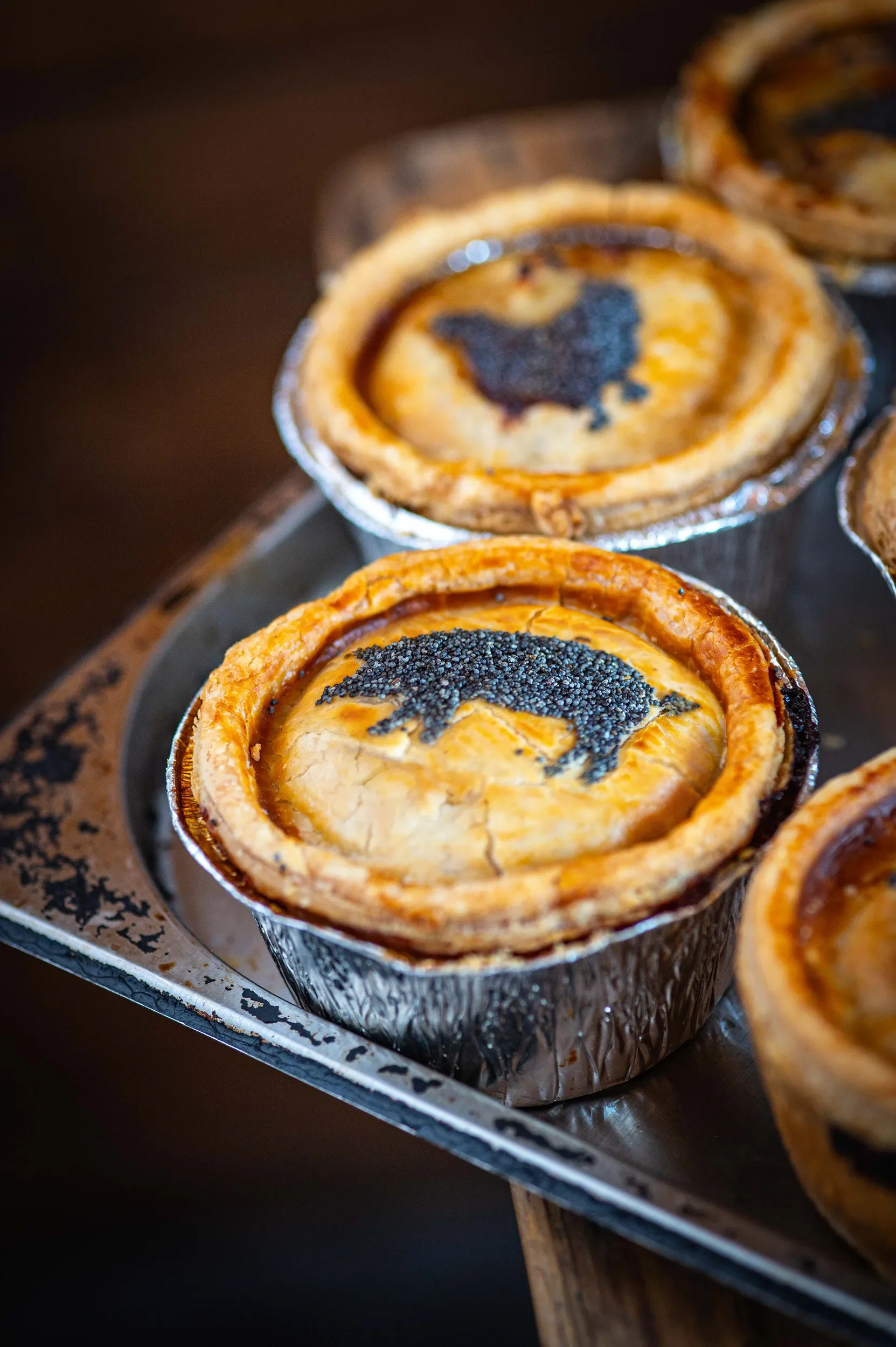 Freshly baked individual meat pies in foil tins on a metal tray, showcasing a distinct pig-shaped poppy seed design on the golden pastry.