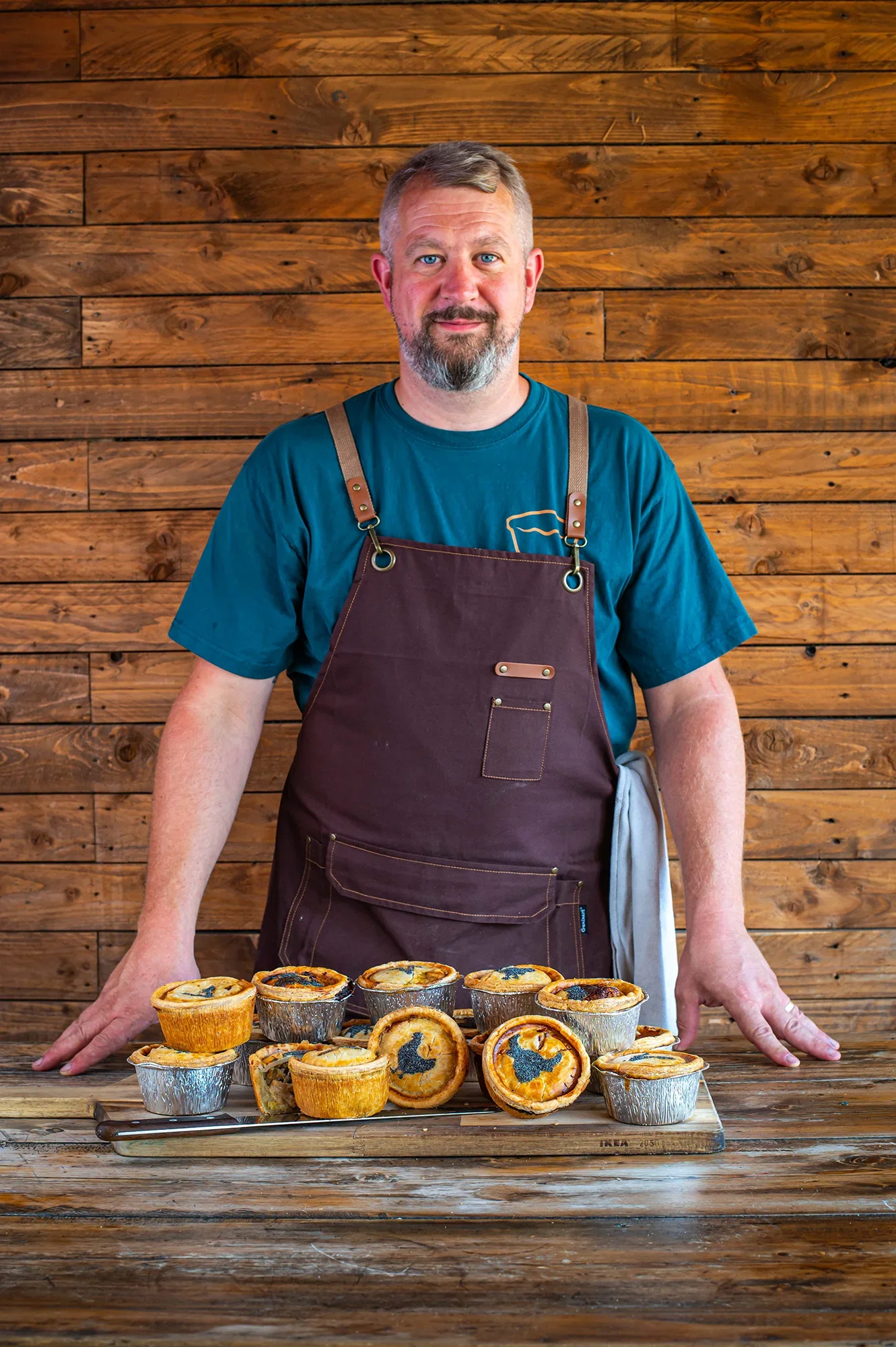 Portrait of Phil, owner and baker of Bristol Pie Society, wearing an apron behind a wooden board stacked with handmade savory pies against a rustic wood wall.