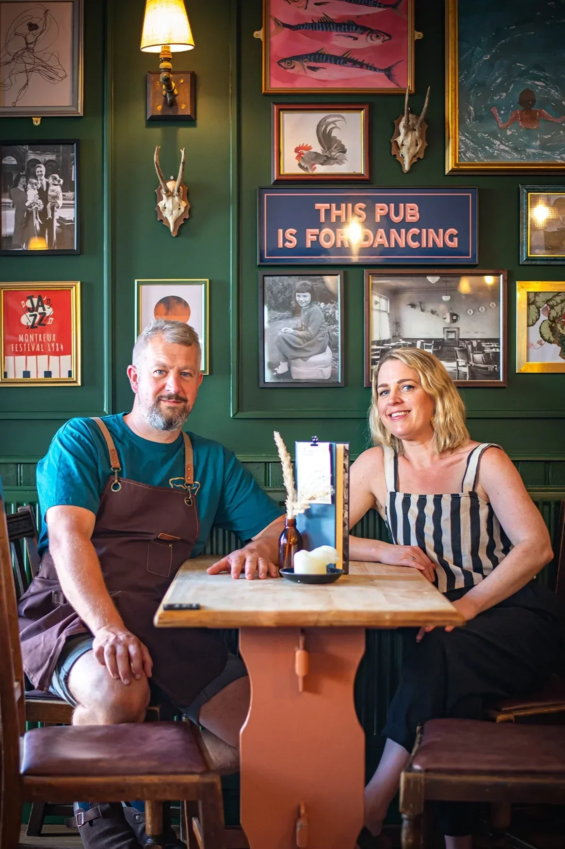 Founders Phil and Amy sitting at a wooden table inside The Gloucester Old Spot pub, in front of a green gallery wall featuring artwork and a sign reading 'This Pub is for Dancing' for Bristol Pie Society.