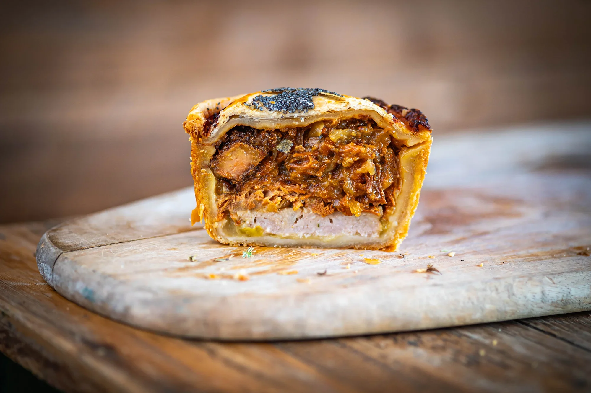 A slice of pie with a flaky crust, filled with seasoned cooked meat and vegetables, topped with a sprinkle of poppy seeds, placed on a rustic wooden cutting board.