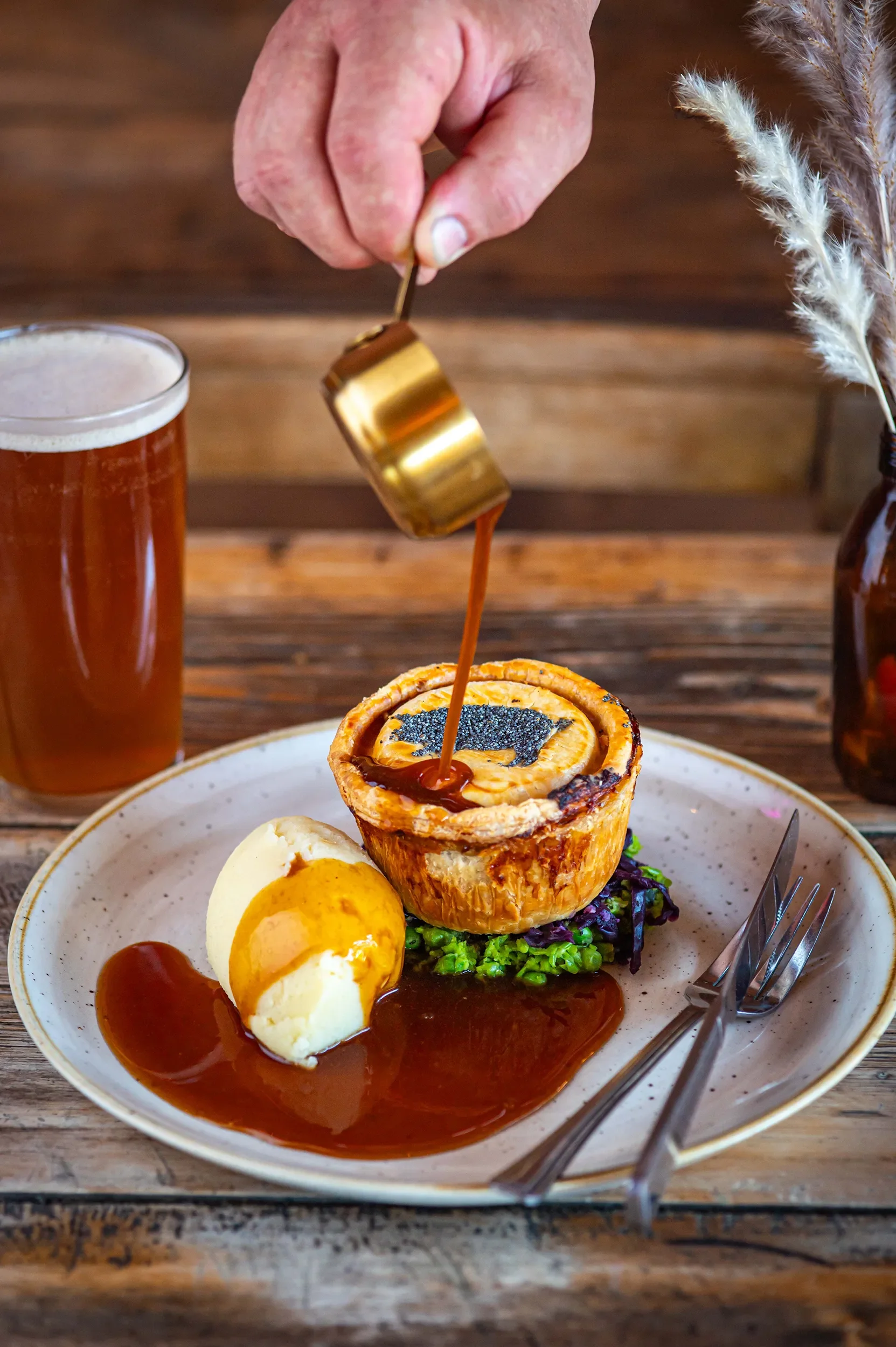 Action shot of thick gravy being poured from a gold ladle over a Bristol Pie Society pie, served with mashed potatoes, mushy peas, red cabbage, and a pint of ale on a rustic wooden table.