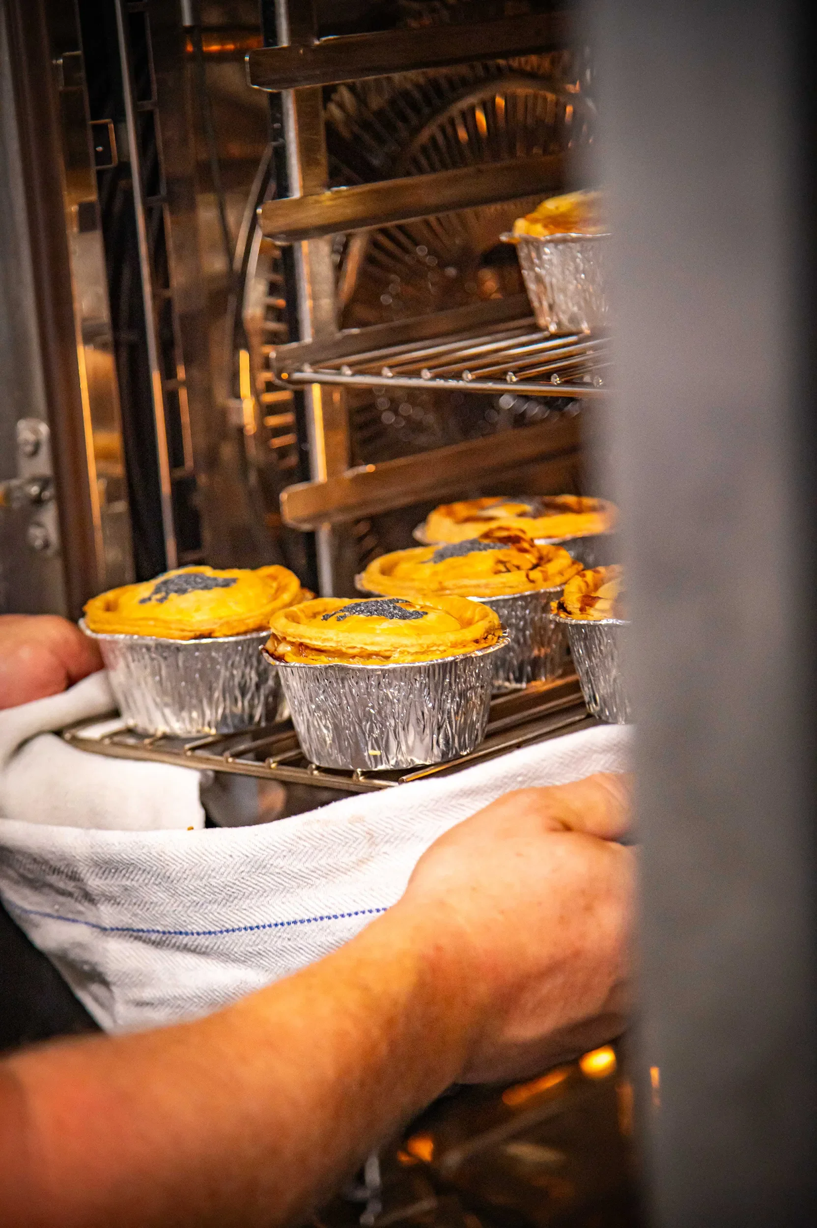 Close-up of a baker using a cloth towel to load a tray of golden, freshly baked savory pies in foil tins into a hot commercial kitchen oven at Bristol Pie Society.