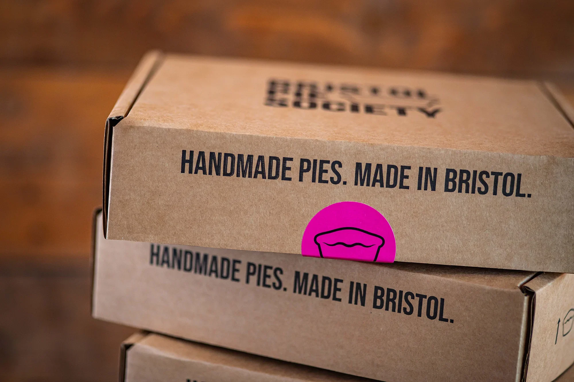 Close-up of stacked cardboard delivery boxes stamped with 'Handmade Pies. Made in Bristol' and a bright pink Bristol Pie Society logo sticker.