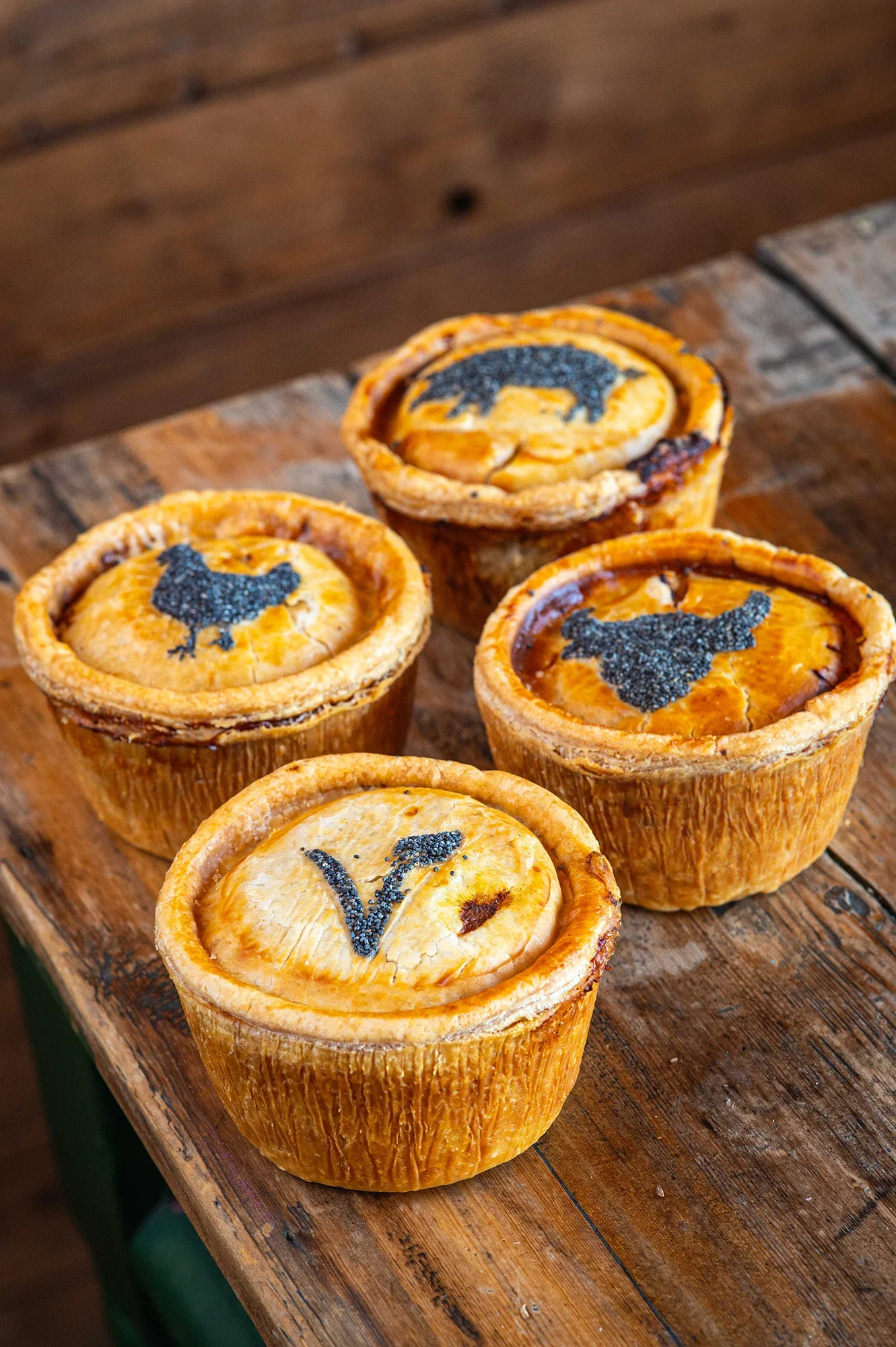 Four golden, handmade Bristol Pie Society pies arranged on a rustic wooden table, featuring poppy seed pastry stencils of a chicken, a pig, a bull, and a vegetarian 'V' symbol.