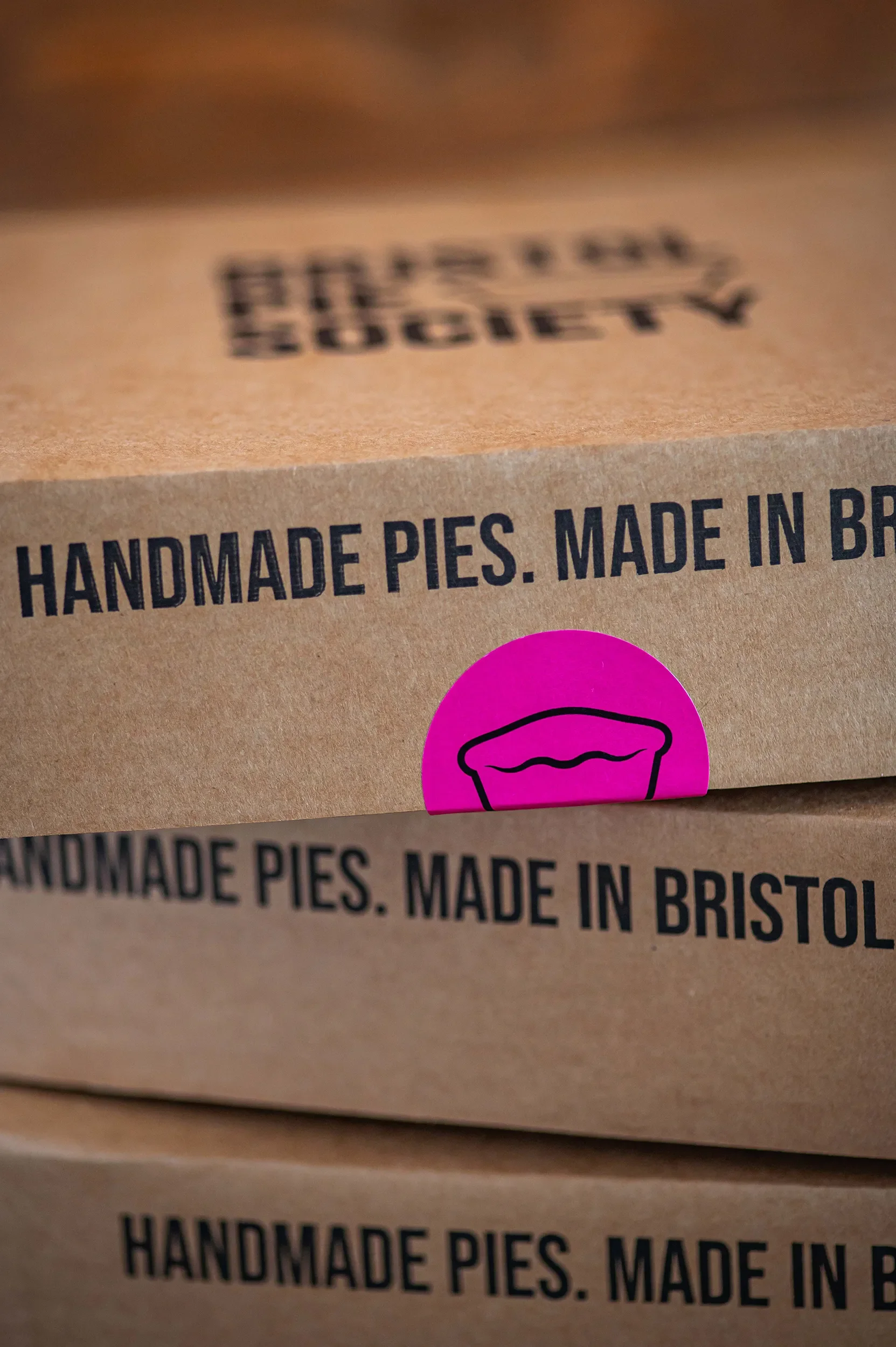 Close-up of stacked cardboard delivery boxes stamped with 'Handmade Pies. Made in Bristol' and a bright pink Bristol Pie Society logo sticker.
