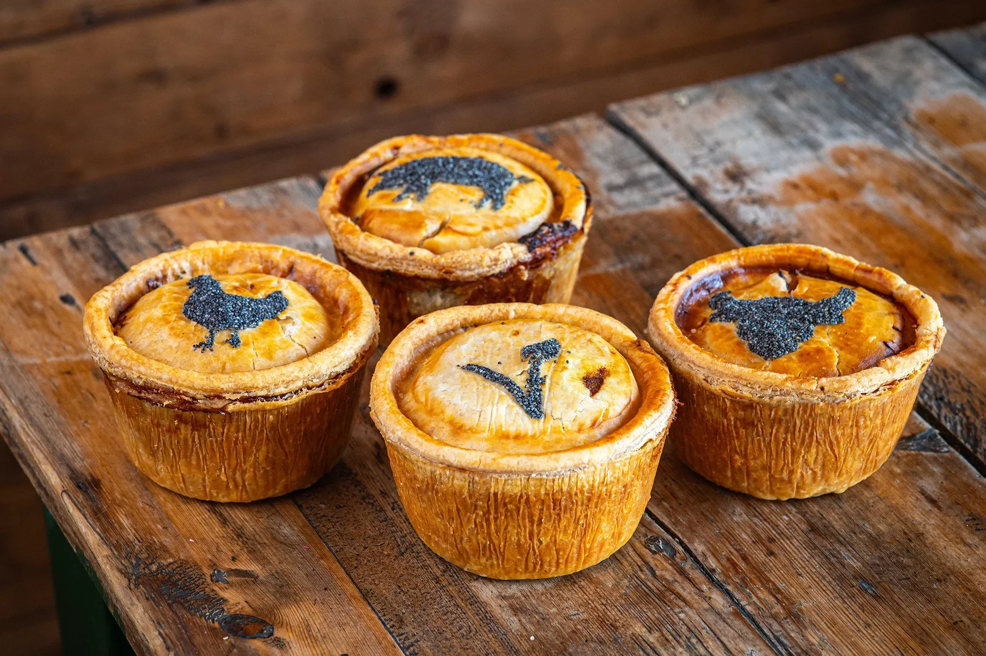 Four golden, handmade Bristol Pie Society pies arranged on a rustic wooden table, featuring poppy seed pastry stencils of a chicken, a pig, a bull, and a vegetarian 'V' symbol.
