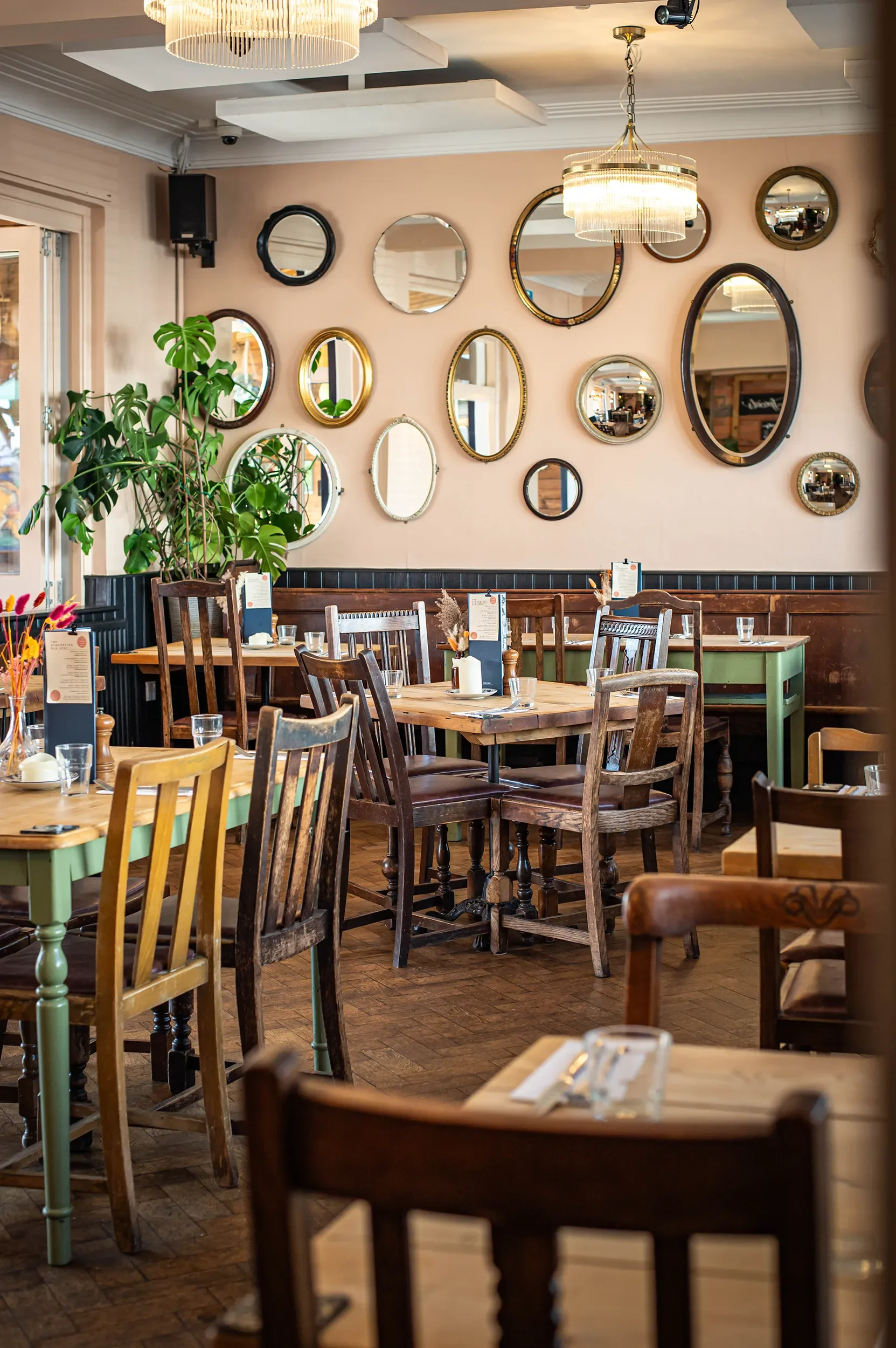 Cosy pub dining area with wooden tables and chairs next to an eclectic green gallery wall with framed pictures and vintage decor.