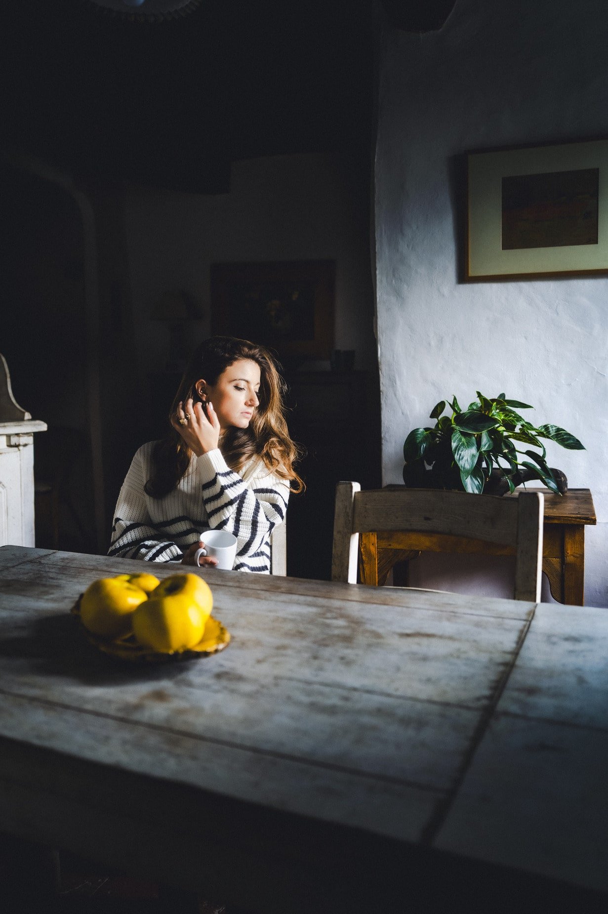 Femme assise à une table en bois, tenant une tasse, avec des citrons sur la table et une plante en arrière-plan.