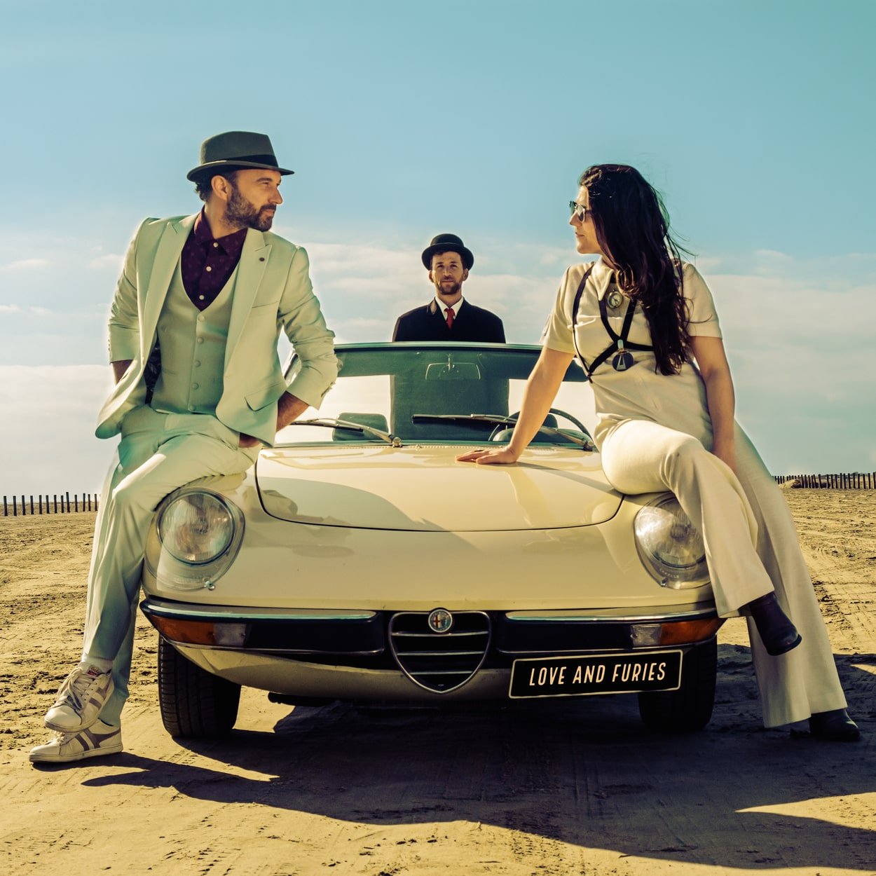 Trois personnes en tenue vintage posent avec une voiture classique sur une plage, sous un ciel clair, avec une plaque "LOVE AND FURIES".