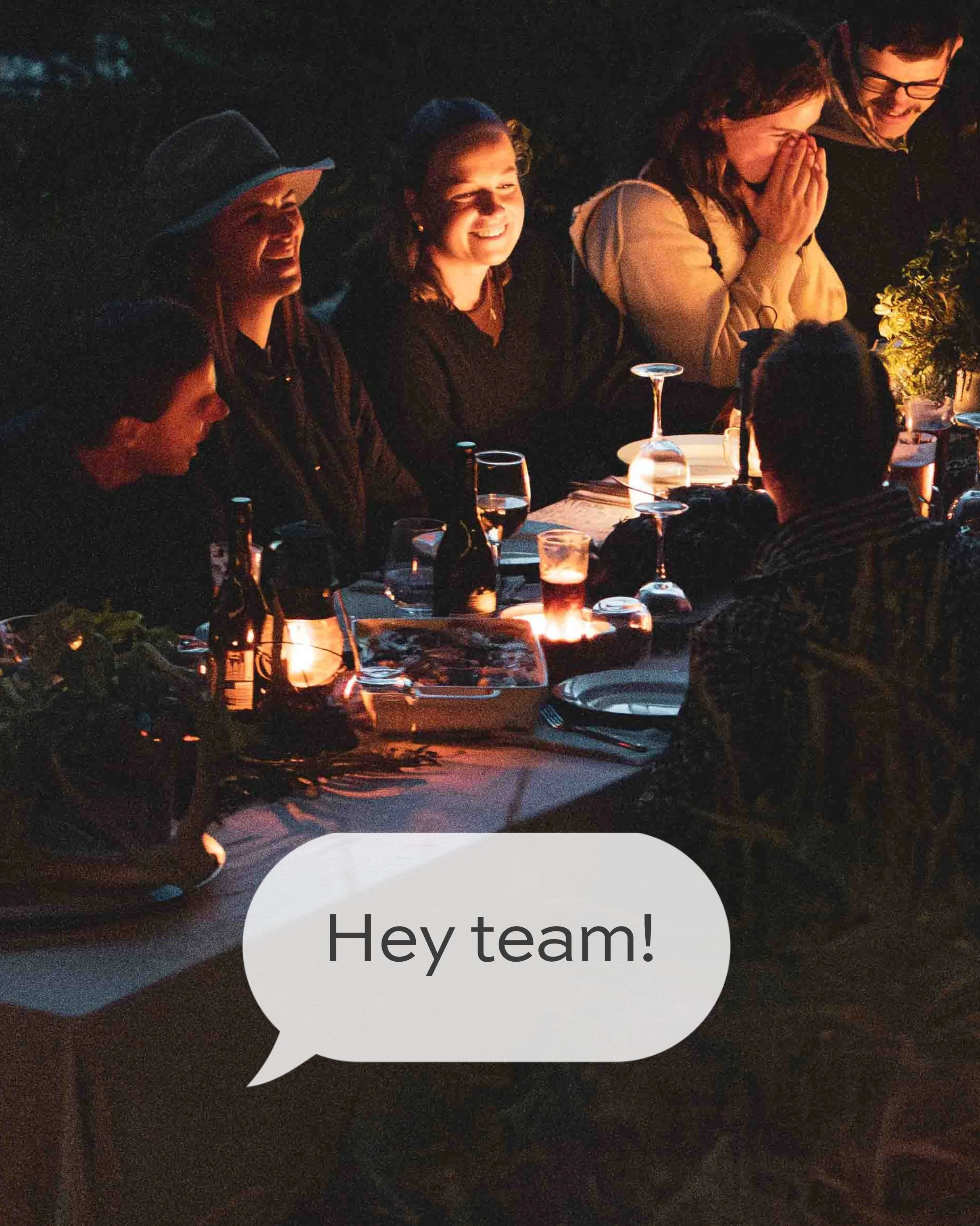 Group of friends sitting around a dinner table at night, smiling and laughing. One person is covering their face with hands, others are in conversation. The scene is warm and festive with dim lighting and drinks on the table.