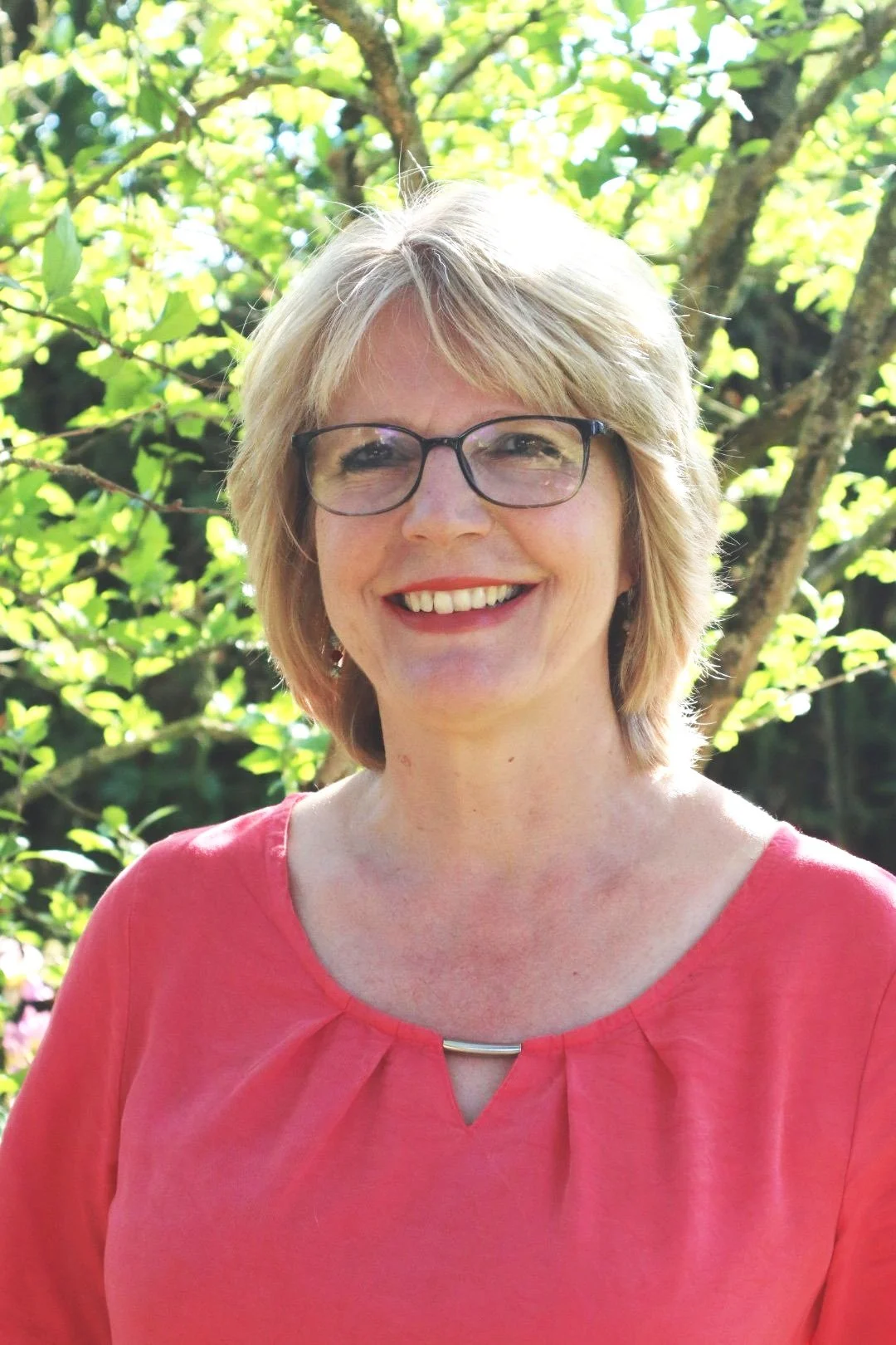 Eine lachende Frau mit Brille und kurzen, blonden Haare vor einem grünen, blättrigen Baum bei sonnigem Wetter.