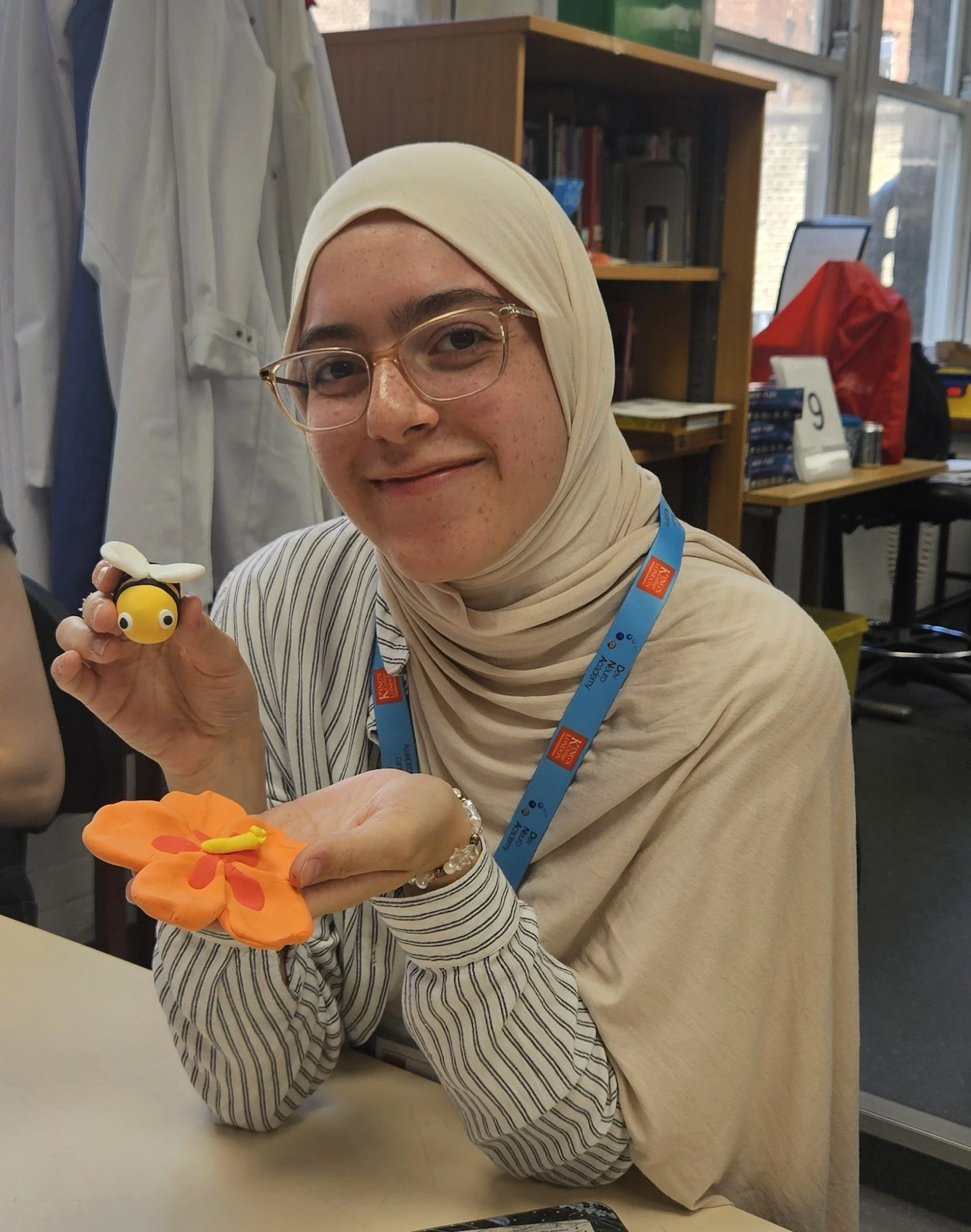 A smiling participant, wearing a cream coloured headscarf and glasses, holds a model of a bee and flower to the camera.