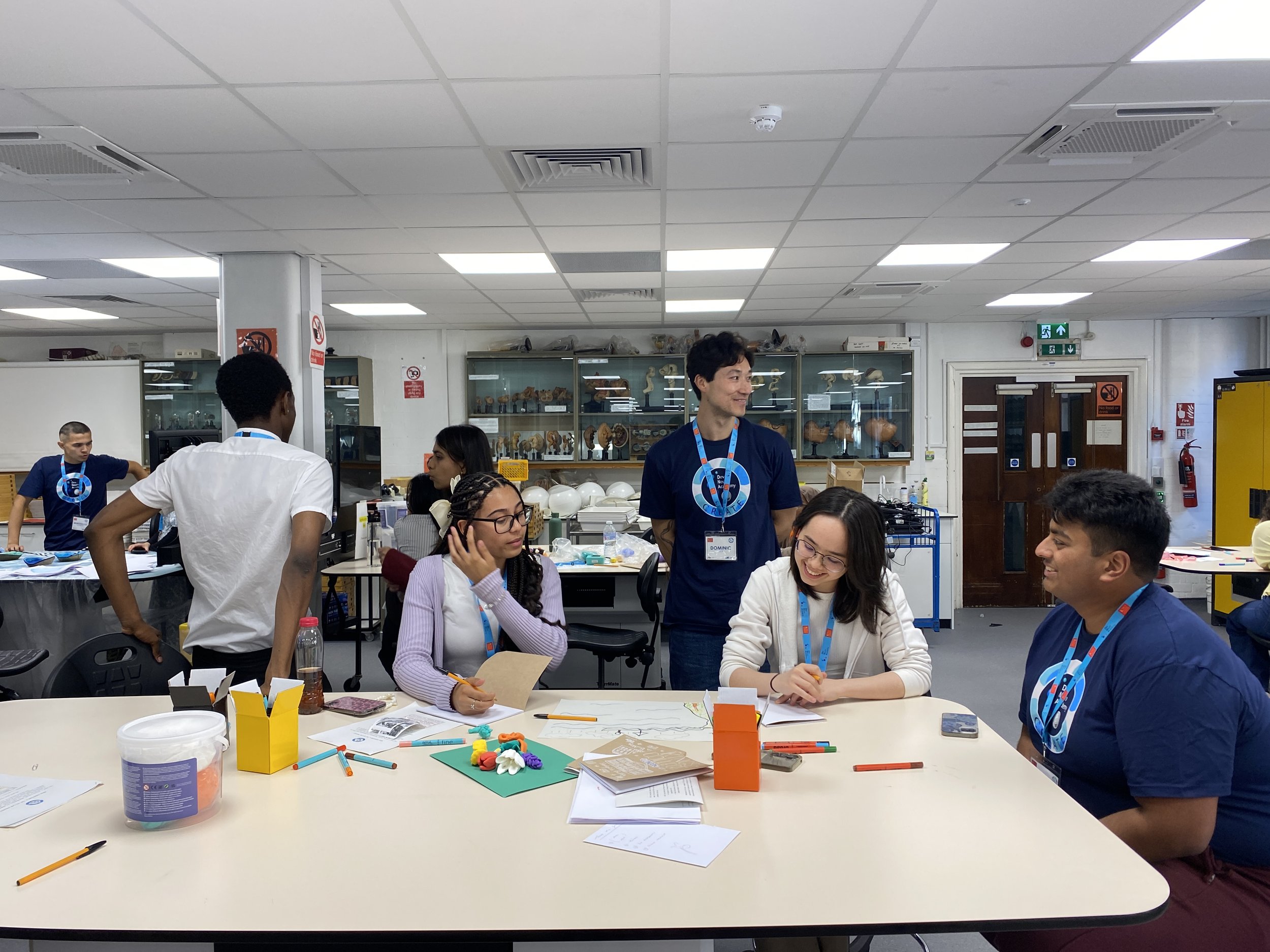 A tutor stands over participants as they draw a signal image on large sheets of paper.
