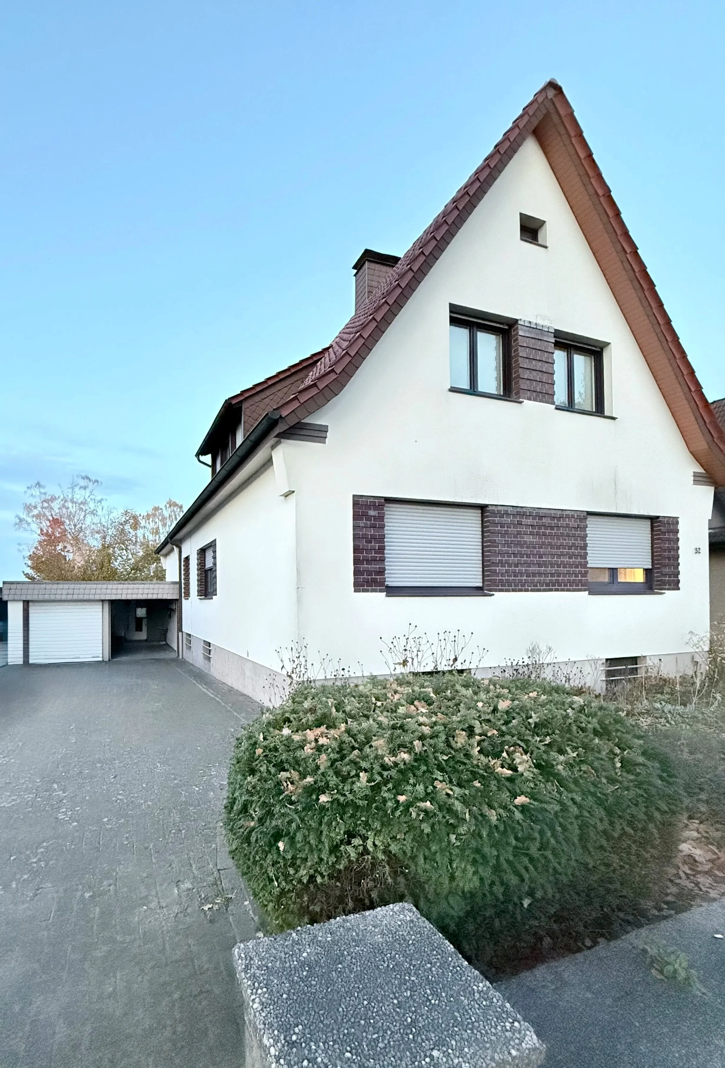 Ein zweistöckiges Haus mit weißer Fassade, braunen Ziegelakzenten und einem roten Satteldach. Es hat mehrere Fenster mit Rollläden und im Vordergrund eine große von Blumen und Sträuchern umgebene Hecke.