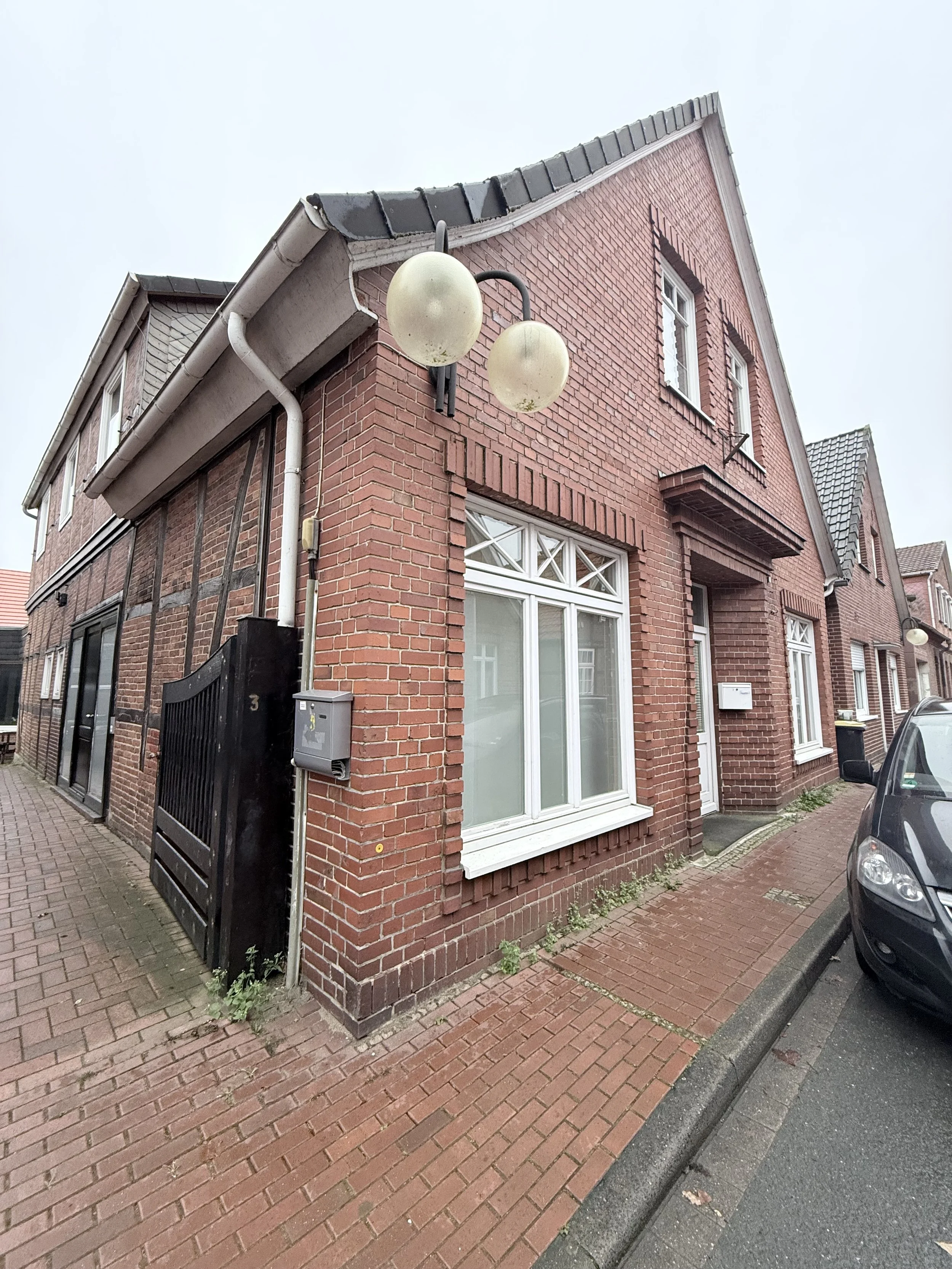 Mehrfamilienhaus aus rotem Ziegelstein mit Doppelhaushälfte, weißen Fenstern und einem schwarzen Tor, auf einem Bürgersteig mit einem geparkten Auto.