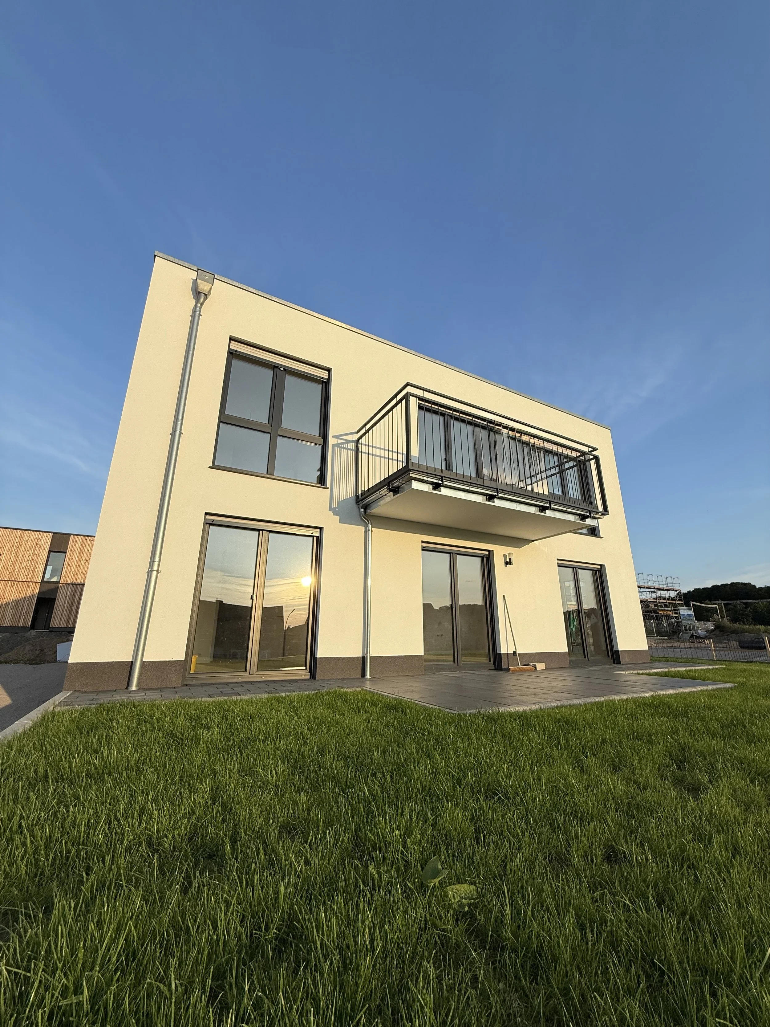Ein modernes weißes Haus mit großen Glastüren und einem Balkon, umgeben von grünem Rasen, bei Sonnenuntergang.