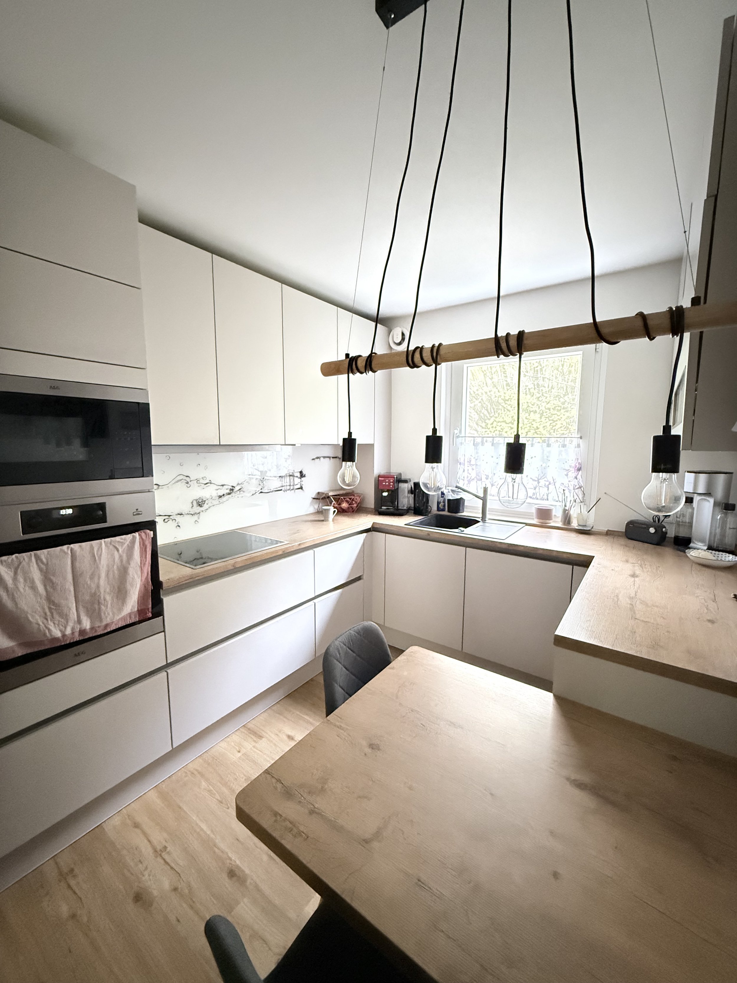 Moderne weiße Küchenschränke, Holzarbeitsflächen und eine Hängelampe aus Glühbirnen über einem Essbereich mit Tisch und Stühlen. Fenster mit Blick nach draußen.