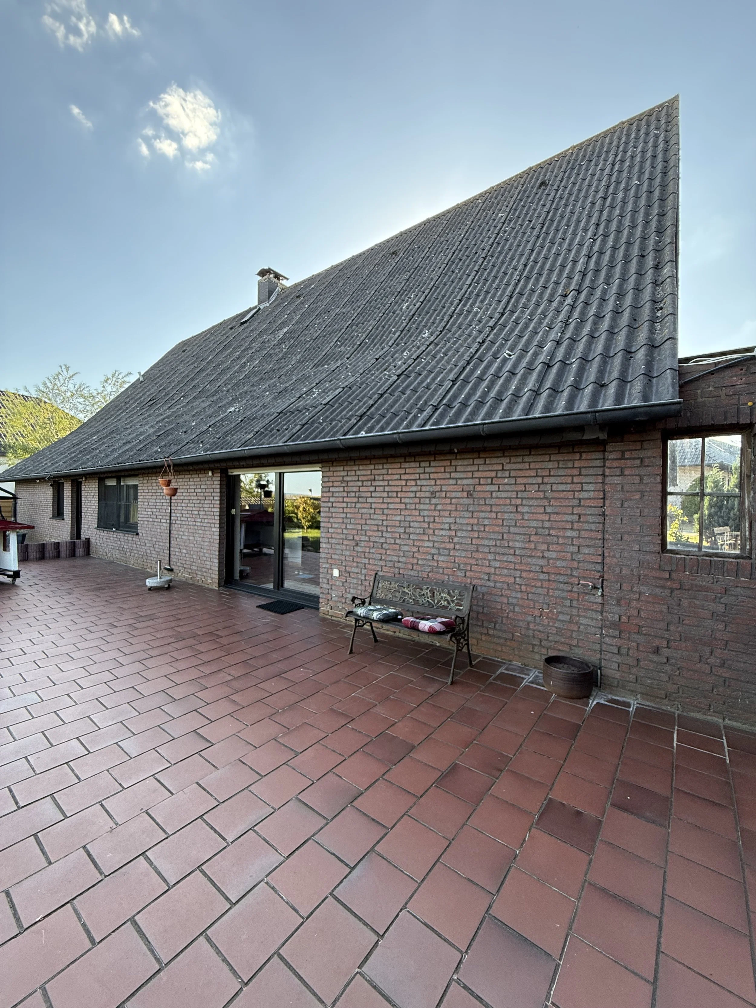 Ein Haus mit einem roten Ziegeldach, die Terrasse ist gefliest und hat eine Bank und einen Blumentopf.