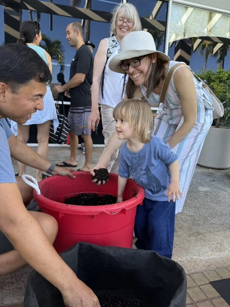 A group of people smiling and engaging in an outdoor activity with a young child, pouring soil into a large red bucket, with a glass building and palm trees in the background.