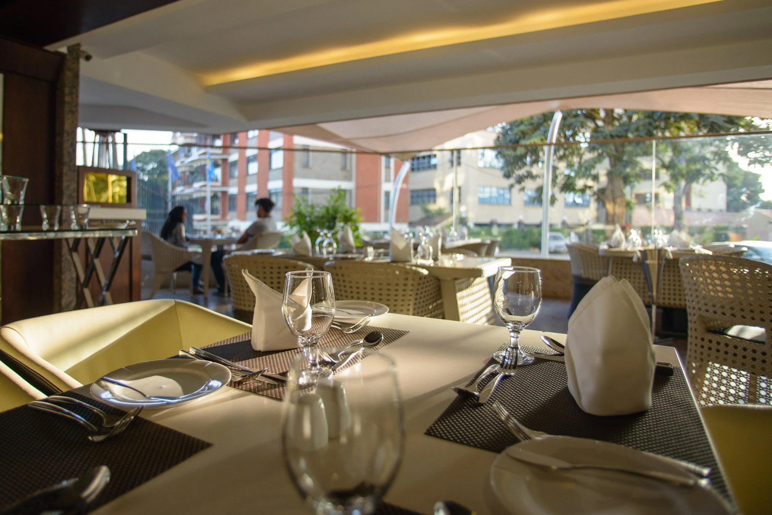 Empty restaurant table set with plates, glasses, silverware, and napkins in a bright, outdoor dining area with other tables and two people seated in the background.