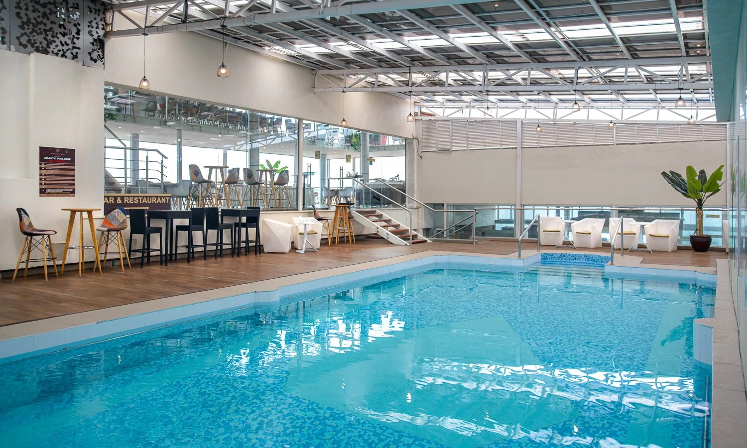 Indoor swimming pool in a modern recreational area with lounge chairs and seating, large windows, and a staircase leading to an upper level.