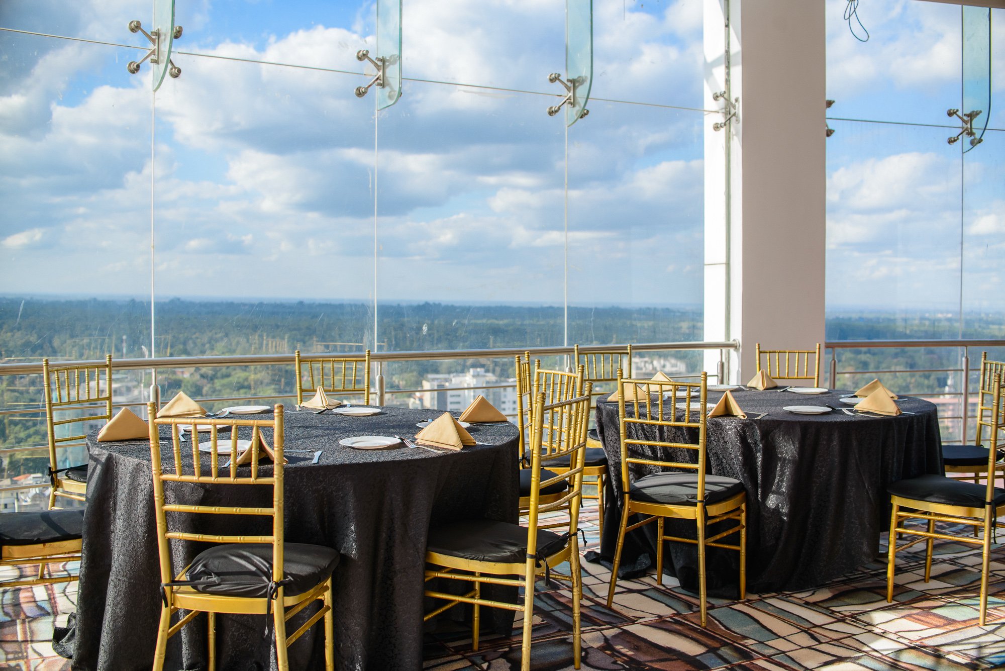 Elegant dining area with round tables covered in black tablecloths, set with white plates and folded beige napkins, surrounded by gold Chiavari chairs, on a high-rise with large glass windows providing a panoramic city and sky view.