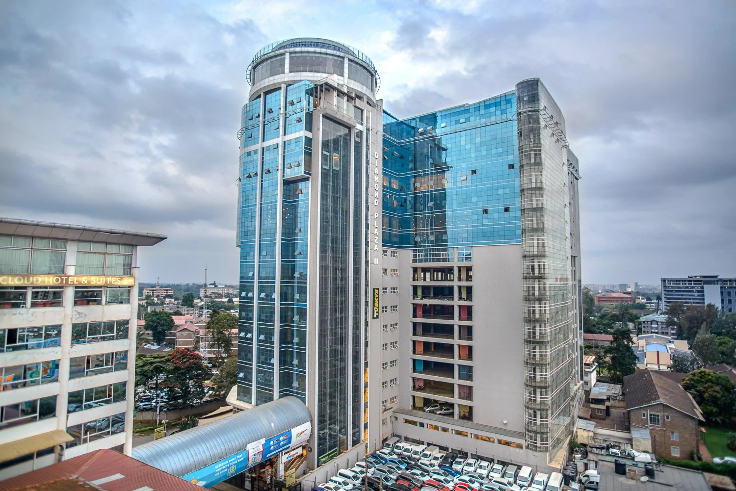 Tall modern glass office building labeled 'Diamond Plaza' on the side with parking lot and surrounding houses below a cloudy sky.