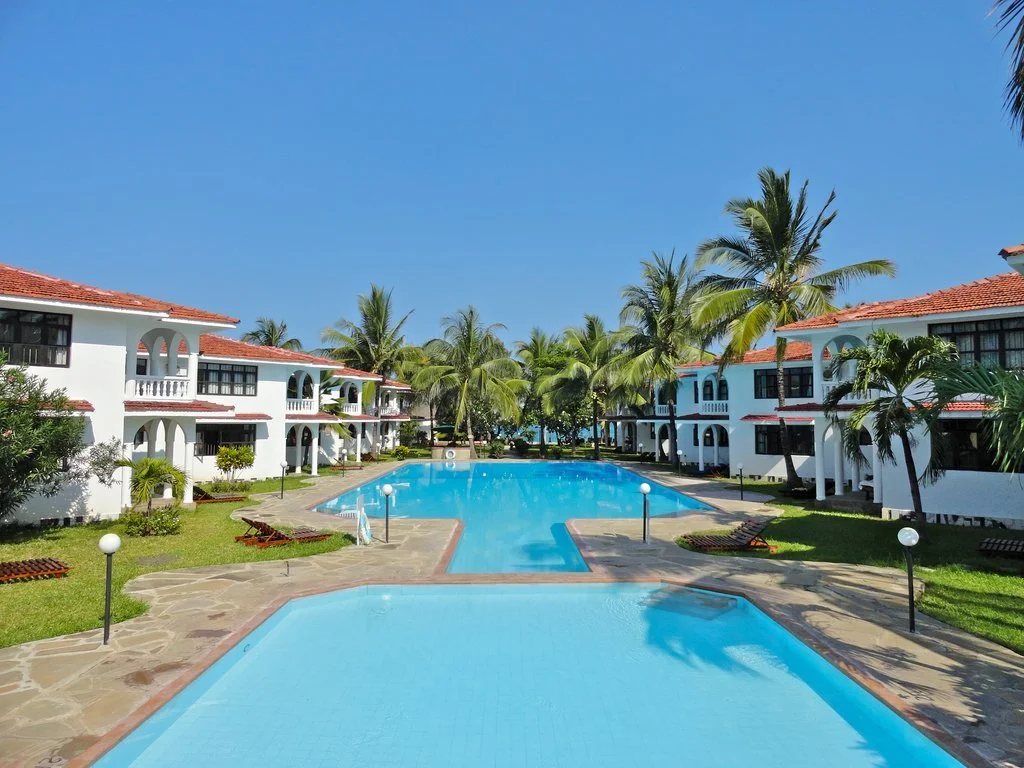 Swimming pool area with white Mediterranean-style buildings, palm trees, and clear blue sky.