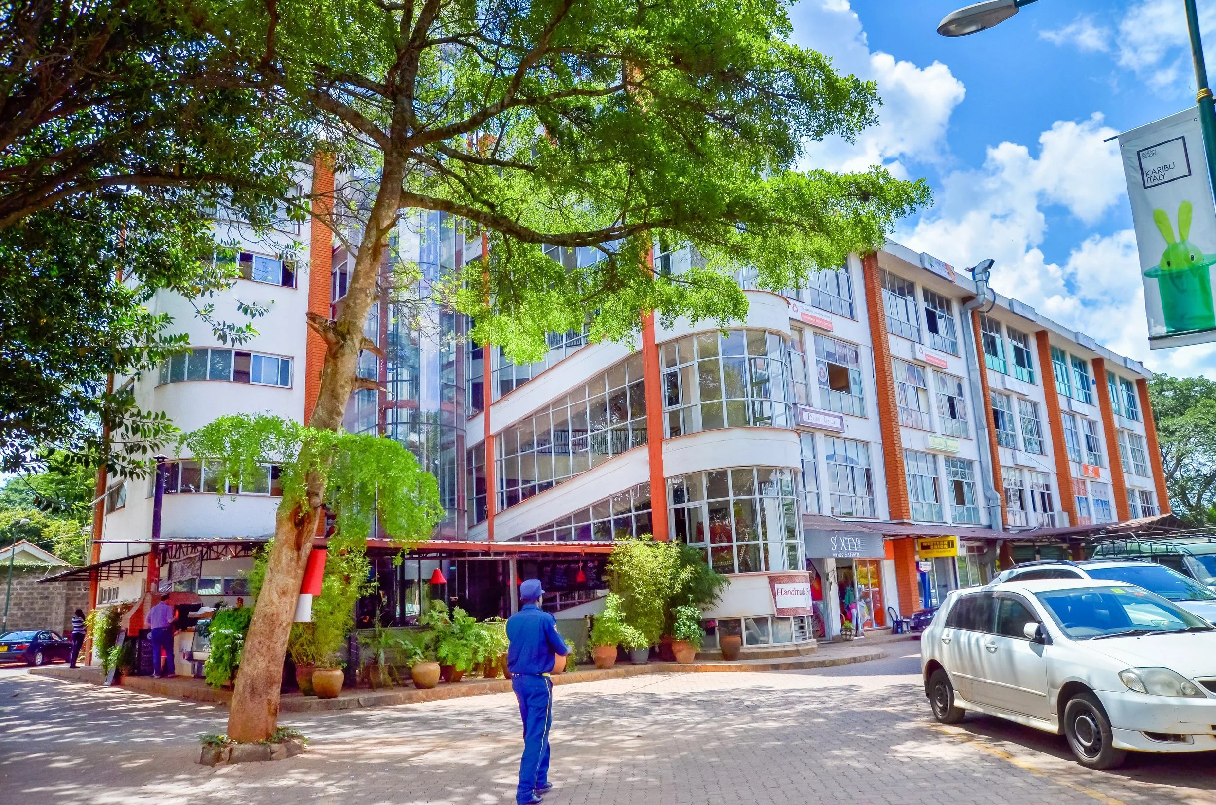 Multi-story building with glass windows, surrounded by trees, with parked cars and a person in blue standing nearby. Clear sky with fluffy clouds.