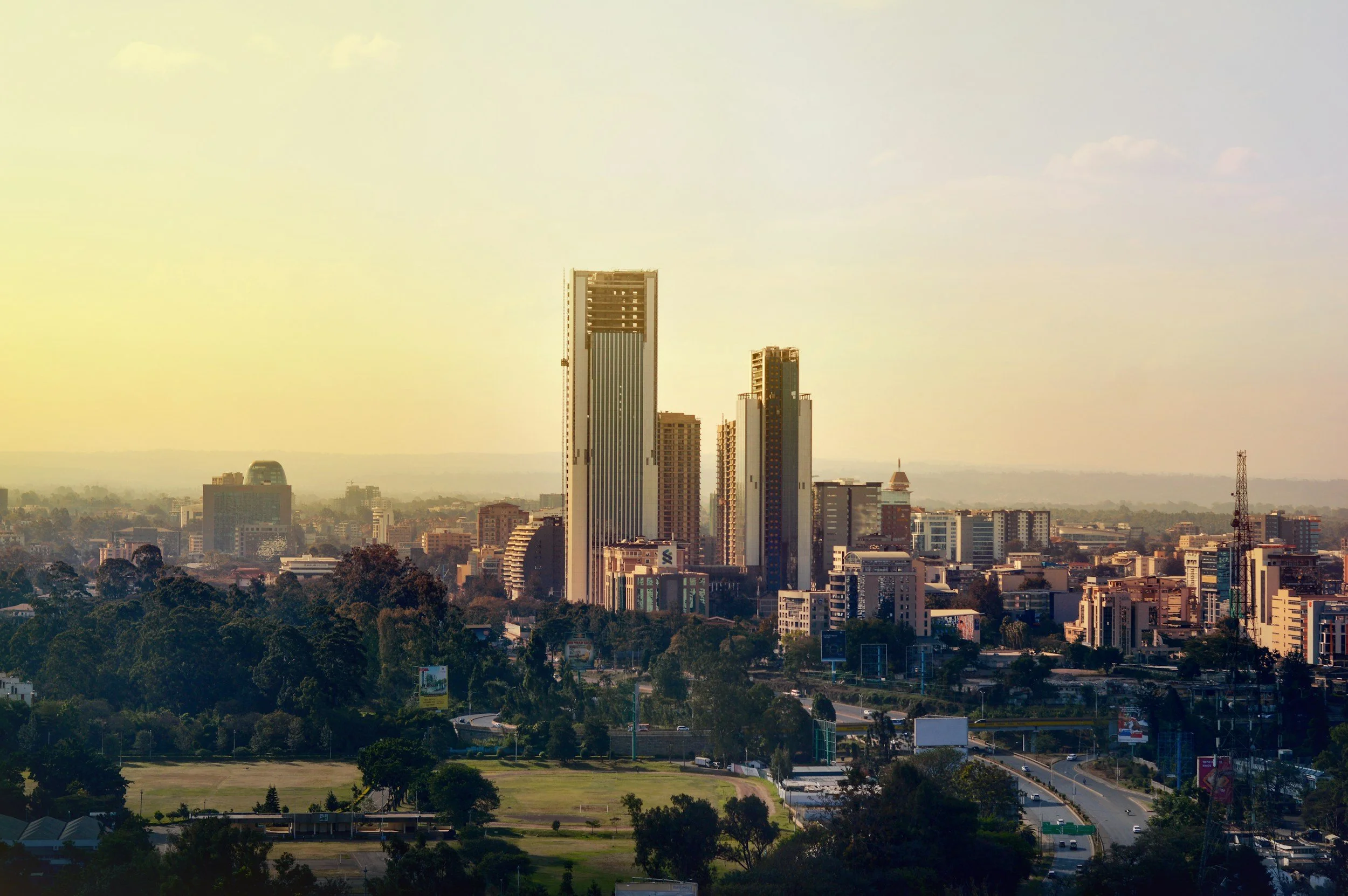 A city skyline with tall modern skyscrapers and a mix of shorter buildings, surrounded by trees and greenery, under a clear sky with some clouds.