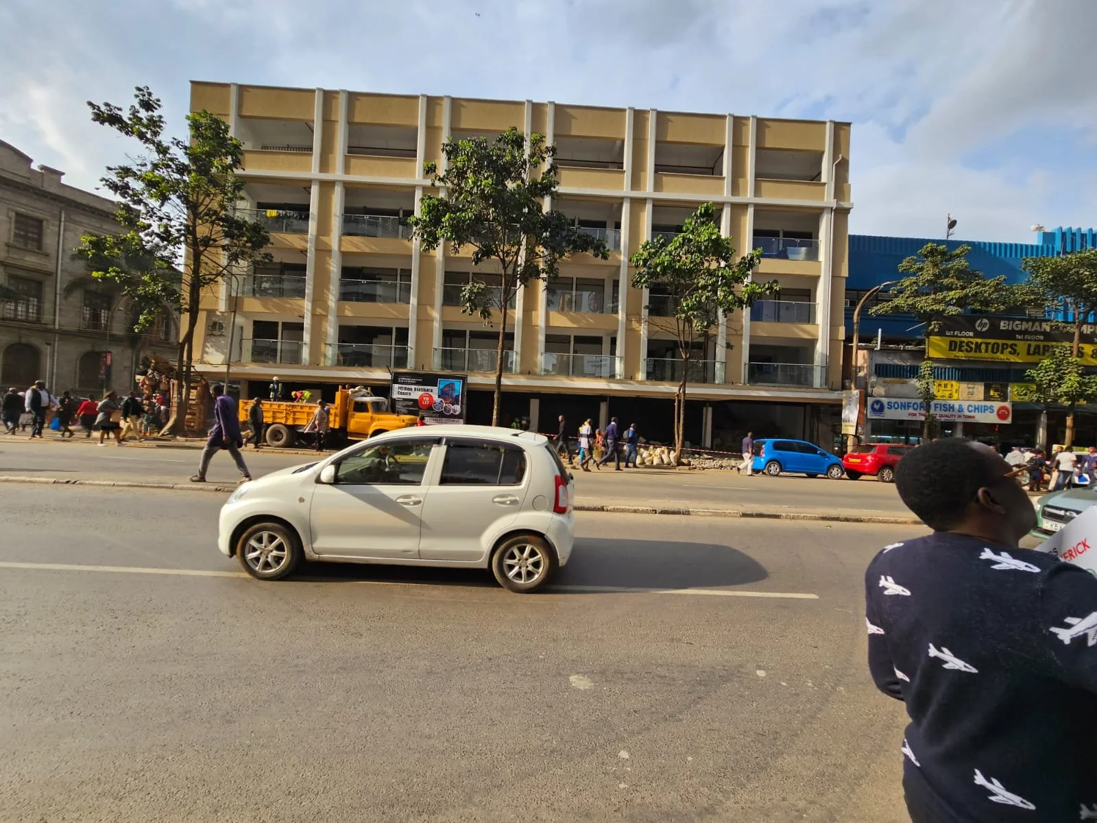 Street scene with a white car in motion in the foreground, pedestrians walking on the sidewalk, a yellow construction vehicle, and a modern multi-story building with trees in front. A person wearing a dark shirt with white patterns is partially visible on the right.