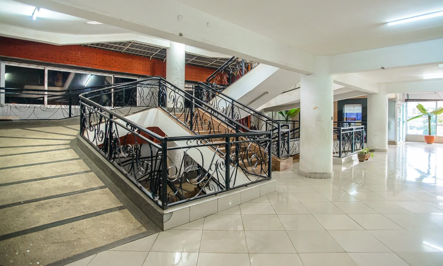 Interior view of a building lobby with curved staircase, black decorative railings, tiled floor, large windows, and potted plants.