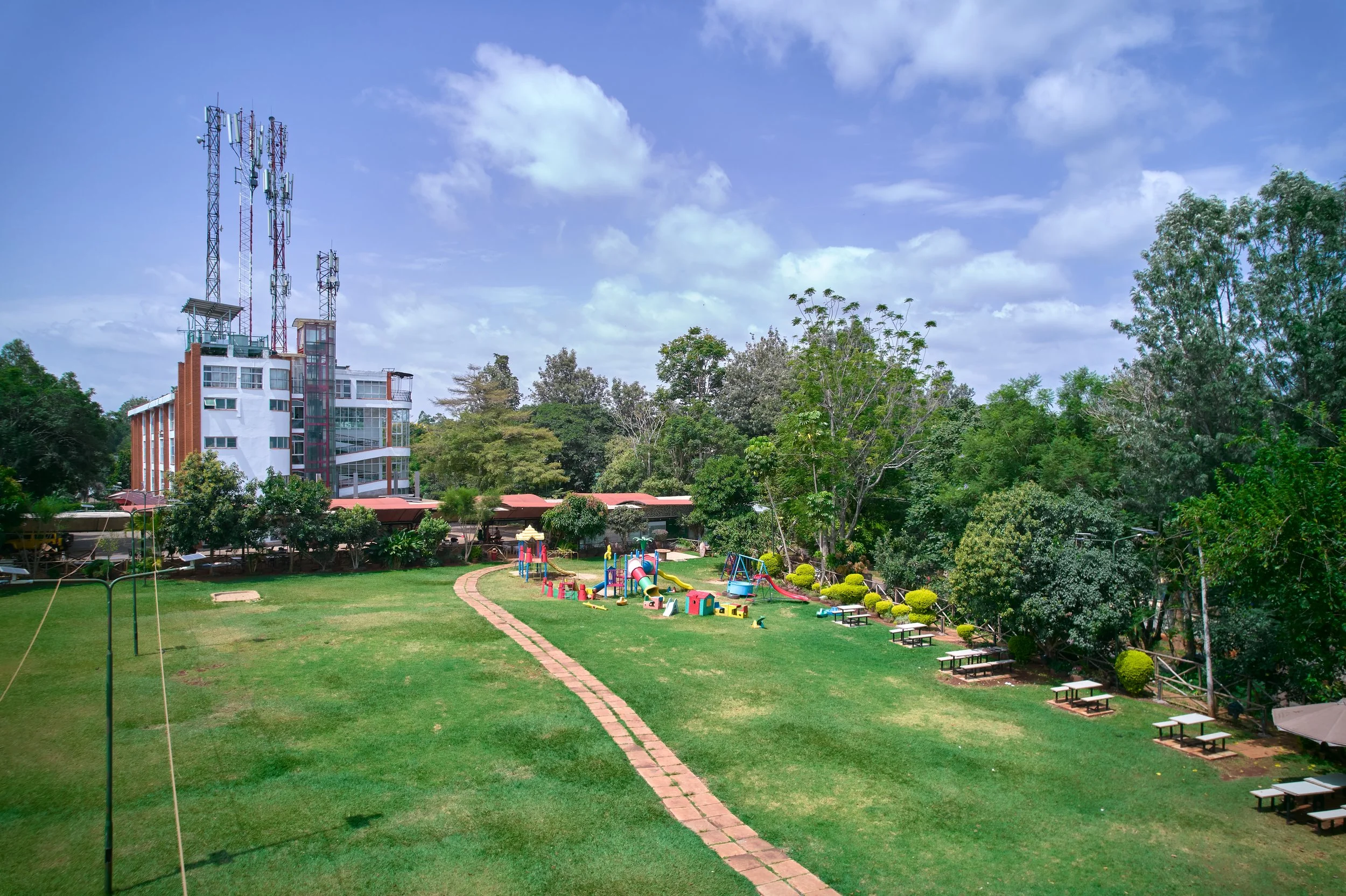 Green lawn with playground equipment, picnic tables, surrounded by trees, under a partly cloudy sky, with a building and communication towers in the background.