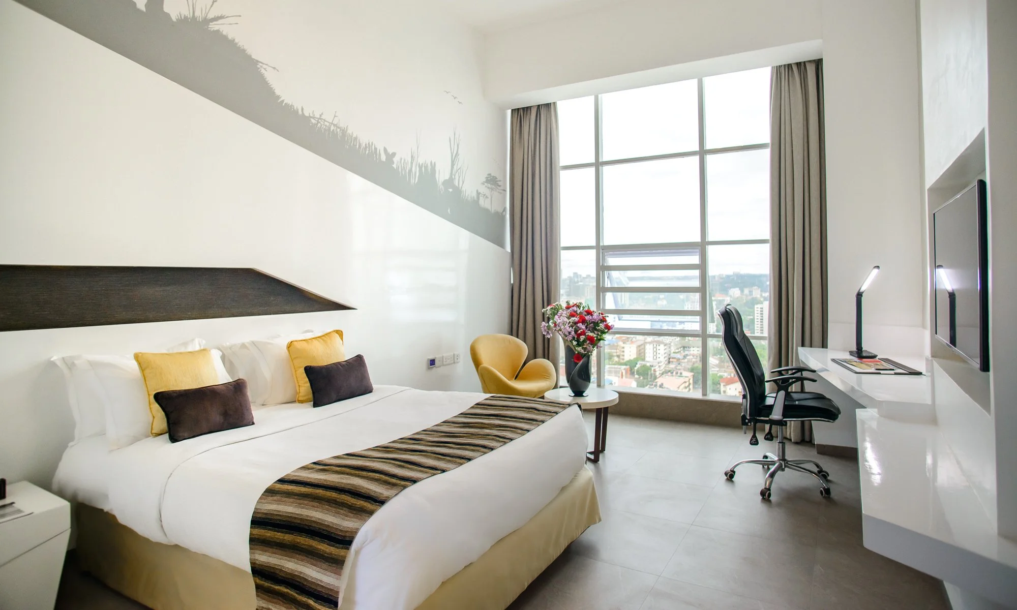 Modern hotel room with large window, bed with yellow and dark pillows, yellow armchair with flowers, and a workspace with a monitor and office chair.