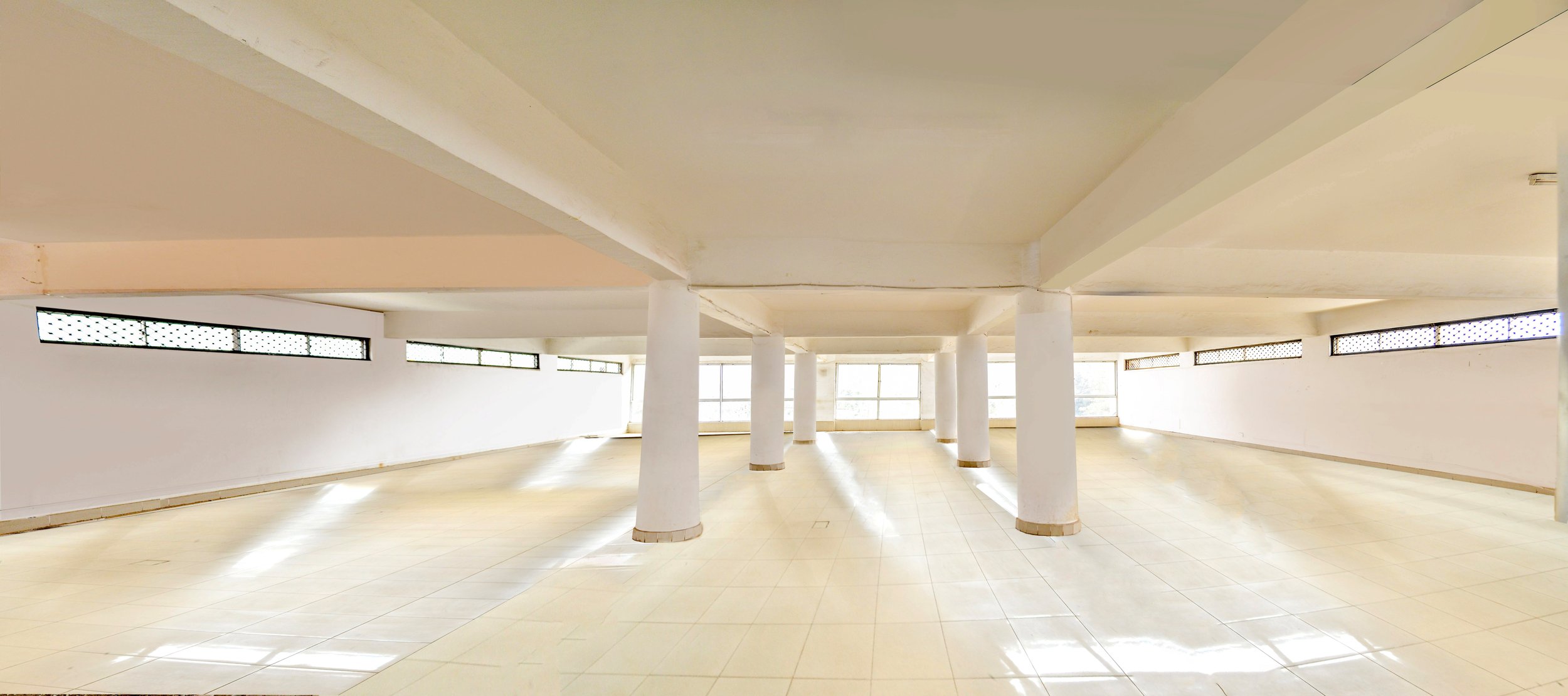 Spacious empty interior room with tiled floor, white walls, multiple small windows near ceiling, and white supporting columns.