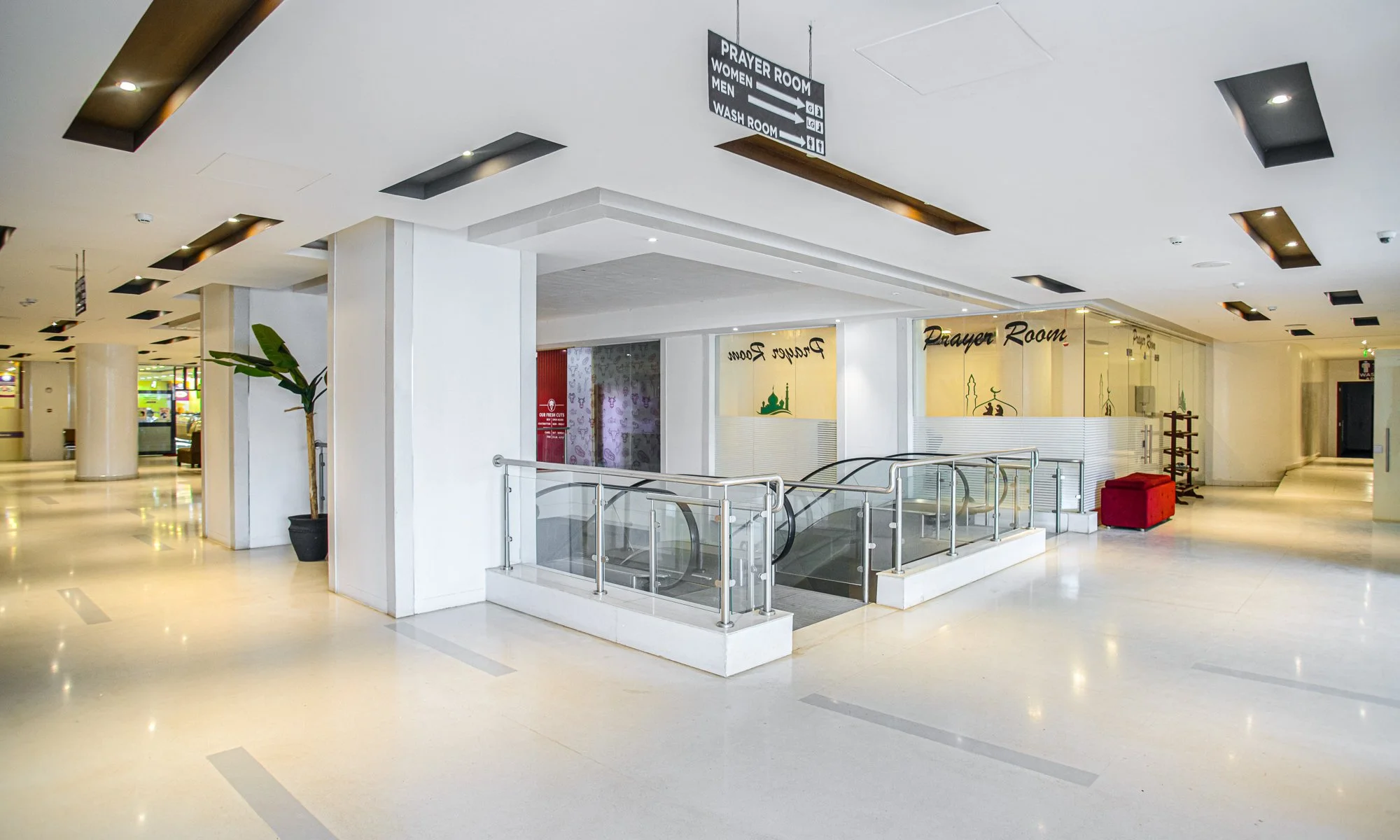 Empty shopping mall corridor with escalator leading down, potted plant, and prayer room signs on the wall.