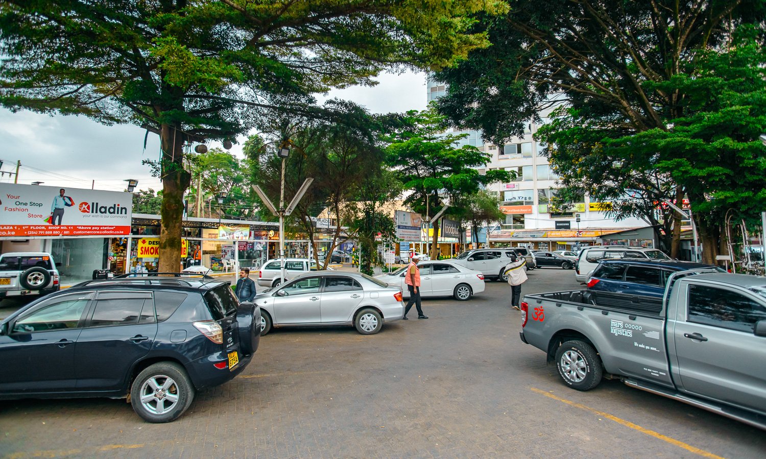 Parking lot with several cars and trees in an urban area, storefronts in the background, people walking, and a mix of commercial buildings.