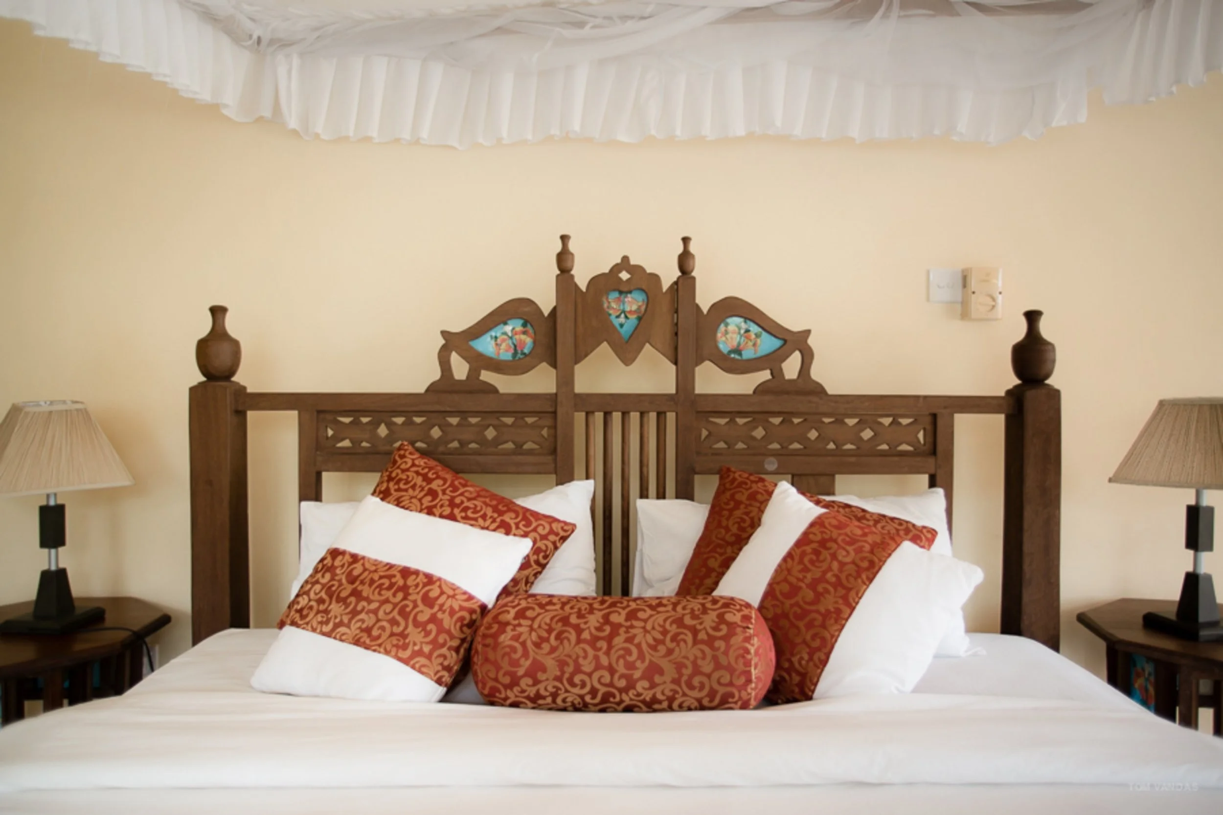 A neatly made bed with a wooden headboard featuring decorative stained glass panels, four pillows with ornate red covers, and a matching cylindrical bolster pillow. There are two lamps on nightstands on either side of the bed, and a white canopy draped overhead. The background wall is light yellow.
