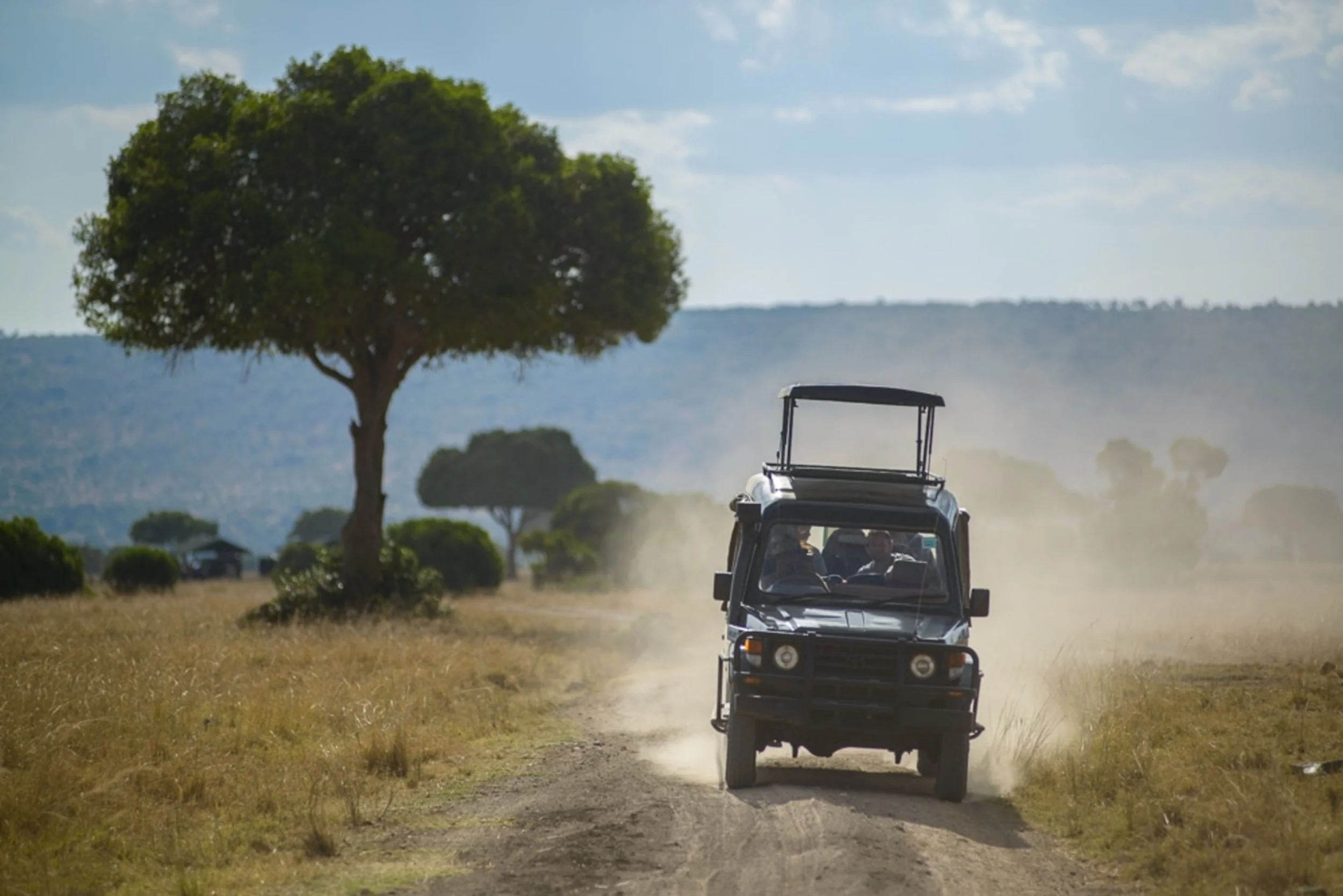 A black utility vehicle driving on a dirt path through a dry grass landscape with scattered trees under a partly cloudy sky.