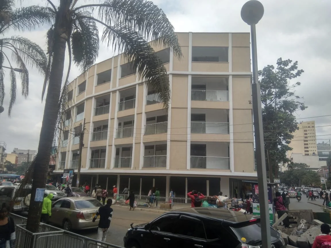 Multi-story commercial building on busy city street, surrounded by cars, pedestrians, street vendors, and palm trees, under cloudy sky.
