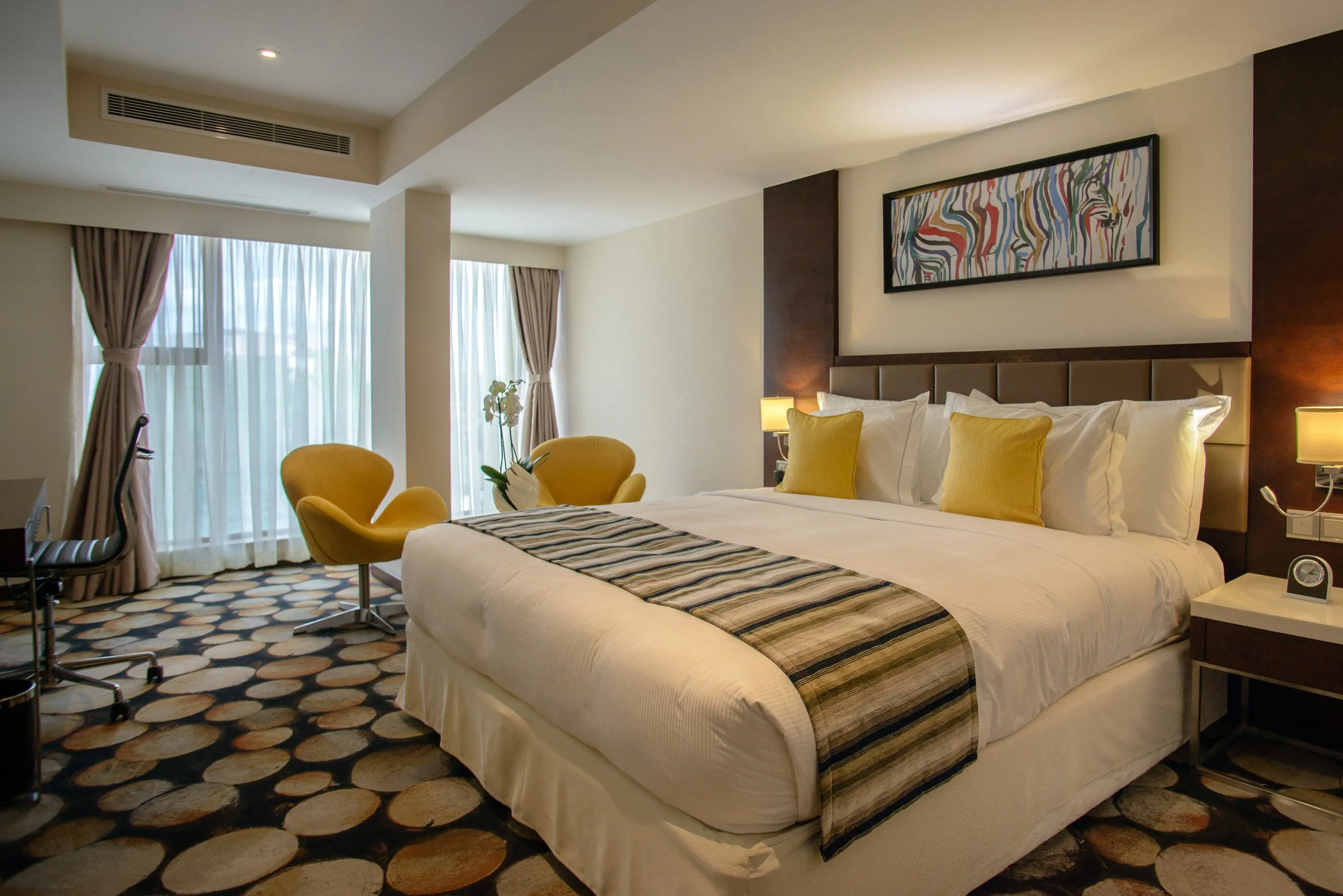 Modern hotel room with a large bed, yellow pillows, and abstract artwork above the headboard. There are yellow chairs, a desk with a black and white striped chair, and large windows with curtains.