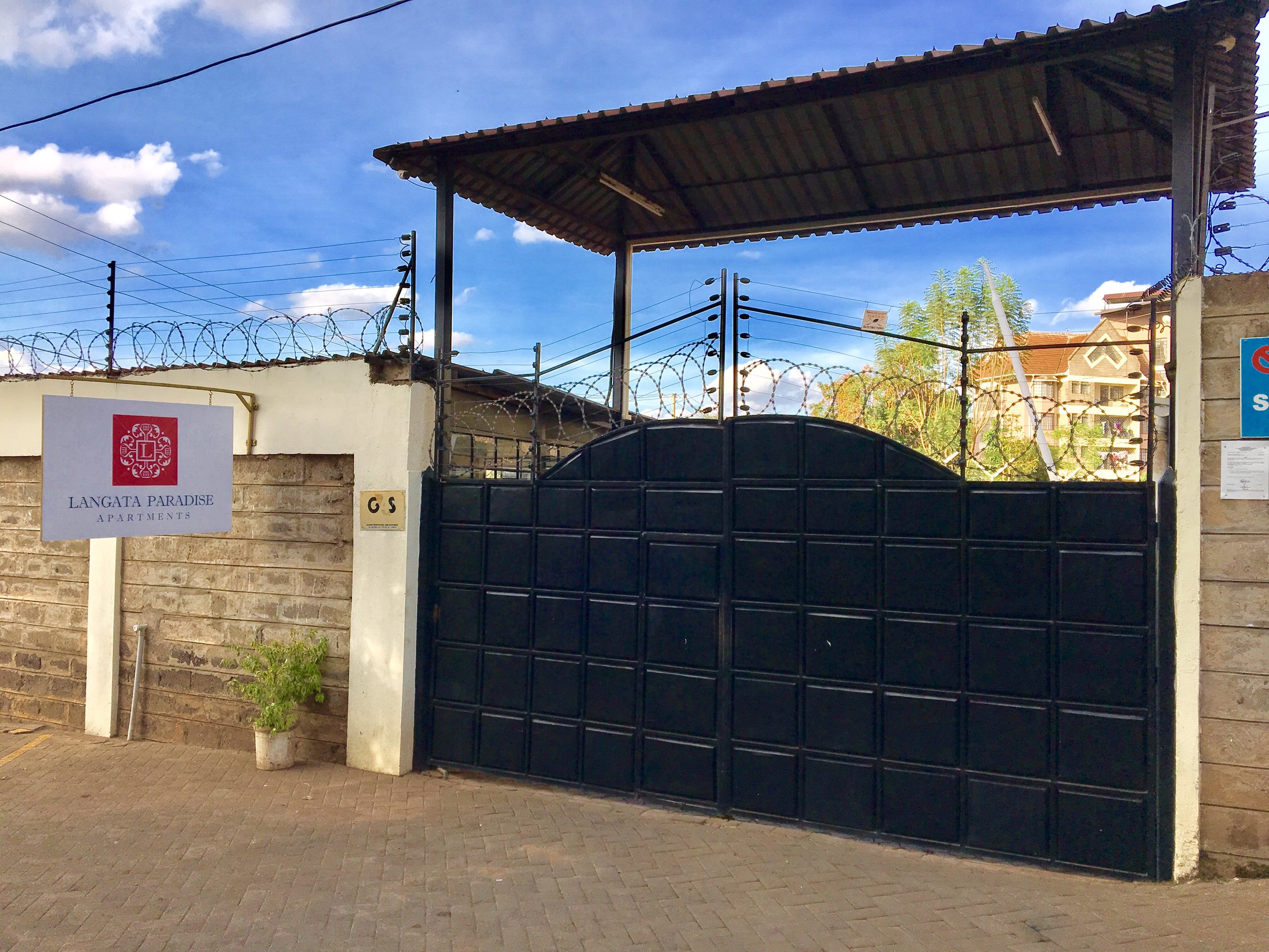 Black metal gate with barbed wire on top, leading into an apartment complex called Langata Paradise Apartments. A white sign with the apartment name is on the left side of the gate, and potted plants are on either side of the entrance. The sky is partly cloudy with some trees and buildings visible in the background.