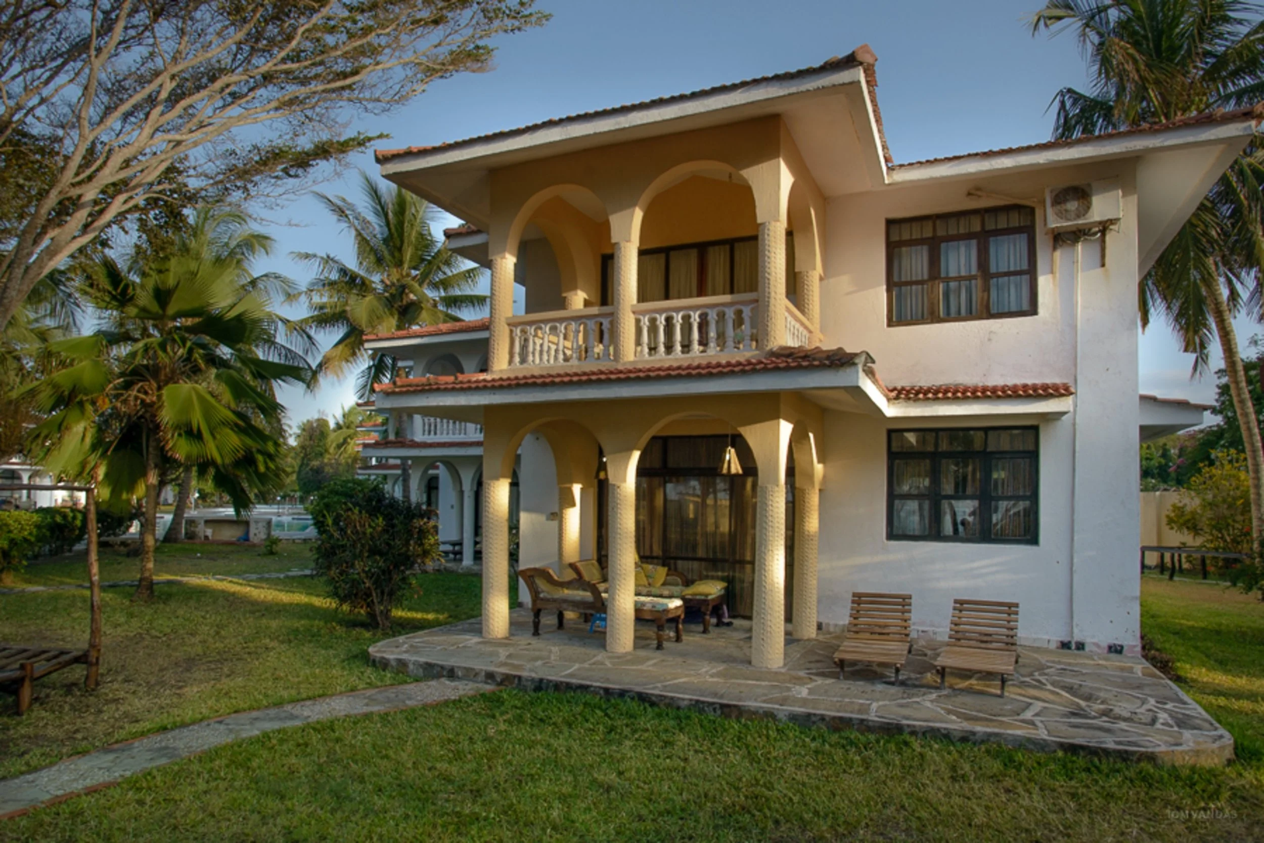 A two-story house with white walls, arched columns, and a red-tiled roof, surrounded by palm trees and greenery, with a patio and outdoor seating area.