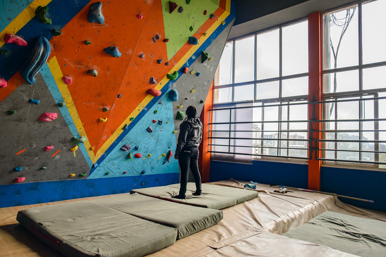 An indoor rock climbing gym with a colorful climbing wall and a person standing on padded mats near the window.