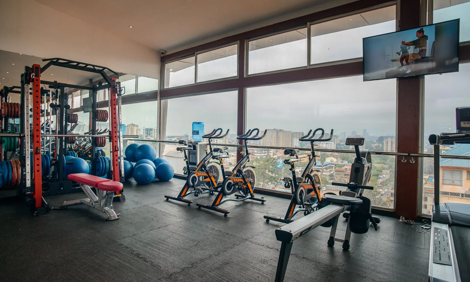 Indoor gym with exercise bikes, weightlifting equipment, stability balls, a treadmill, and a large window with city view.