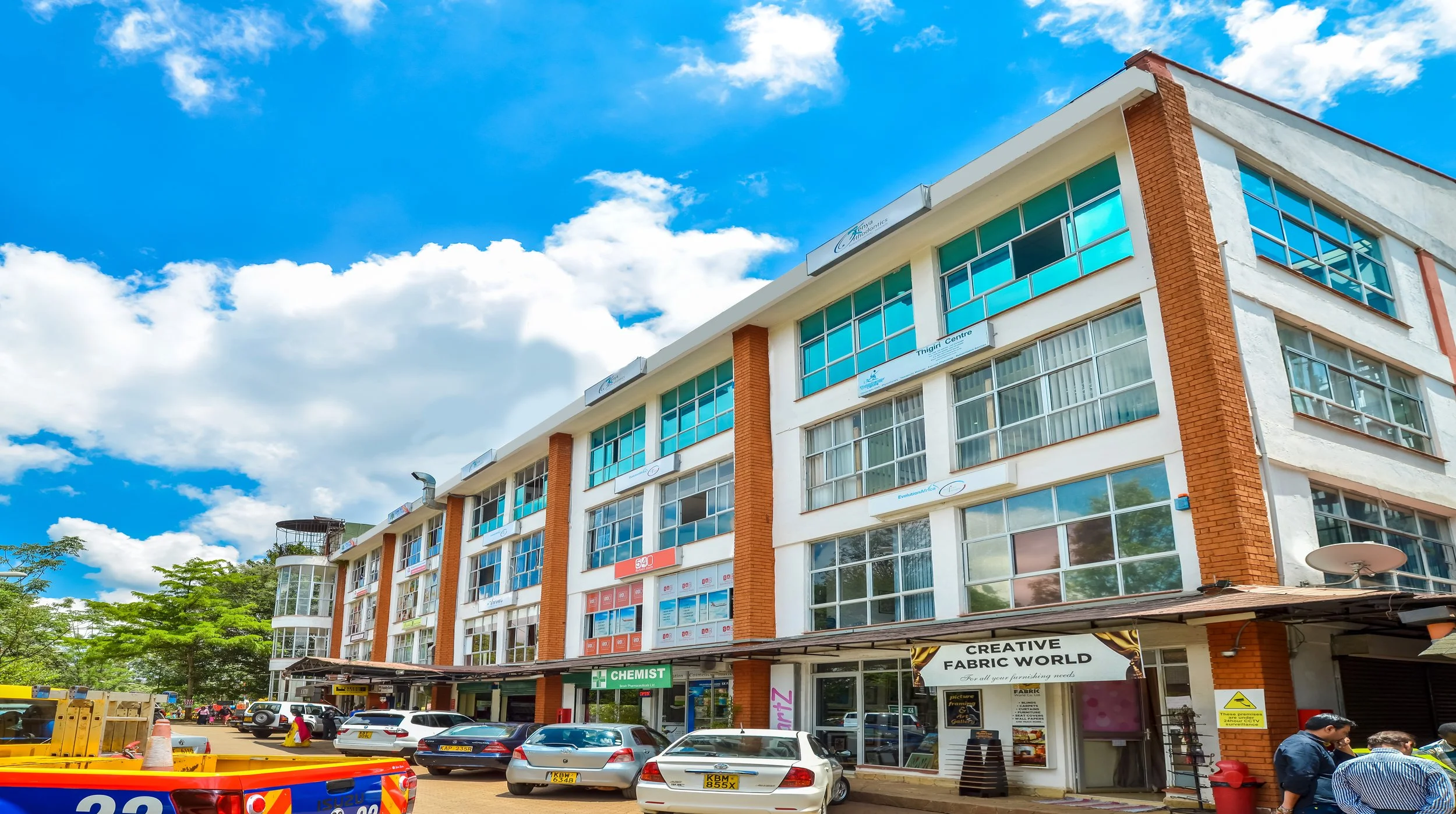 Multi-story commercial building with large glass windows, parked cars in front, trees on the left, and a bright blue sky with clouds.