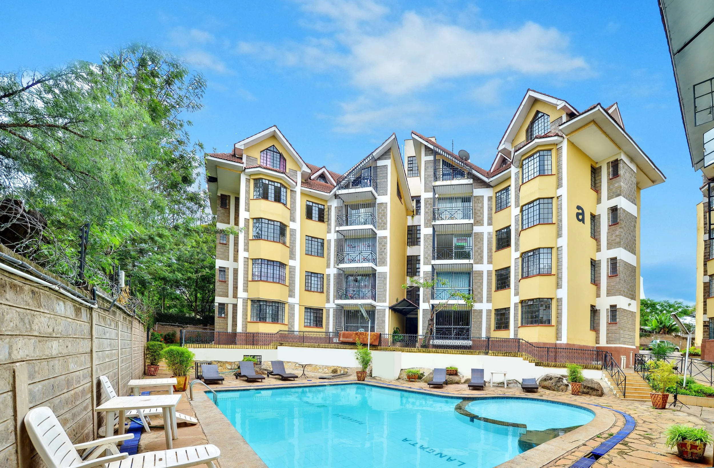 Swimming pool with lounge chairs in front of a yellow apartment building with balconies, under a partly cloudy sky.